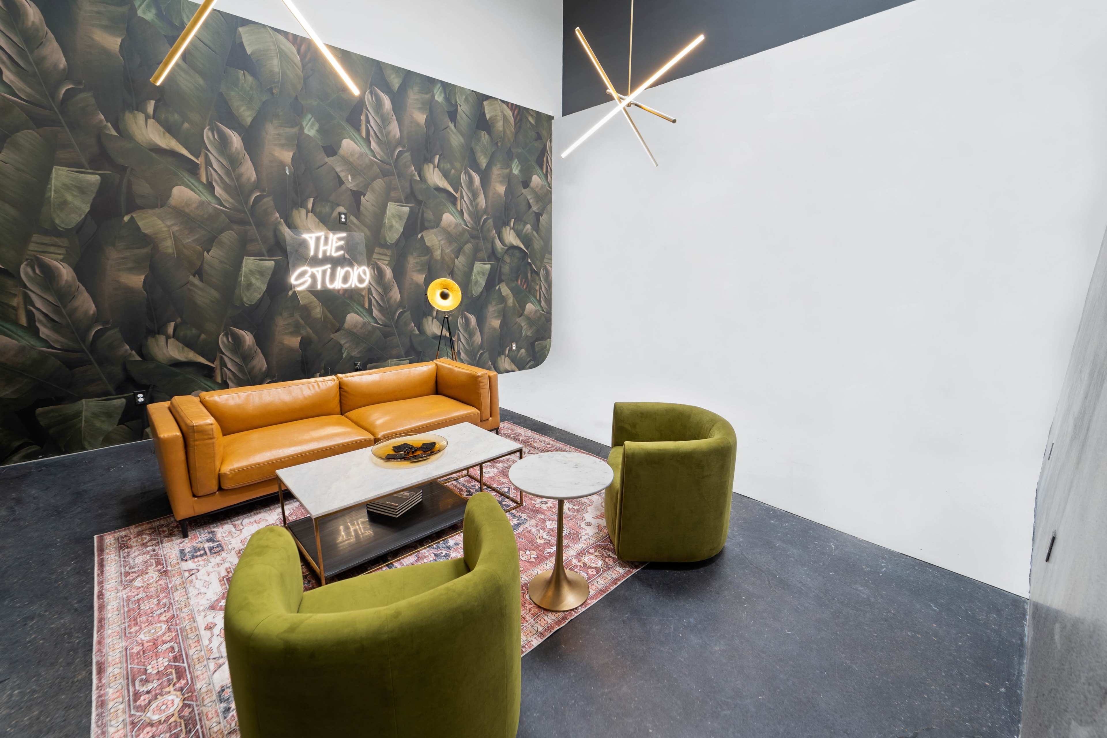 A modern lounge area features a yellow leather sofa, two green velvet chairs, a marble coffee table, and a wall decorated with a leafy mural.