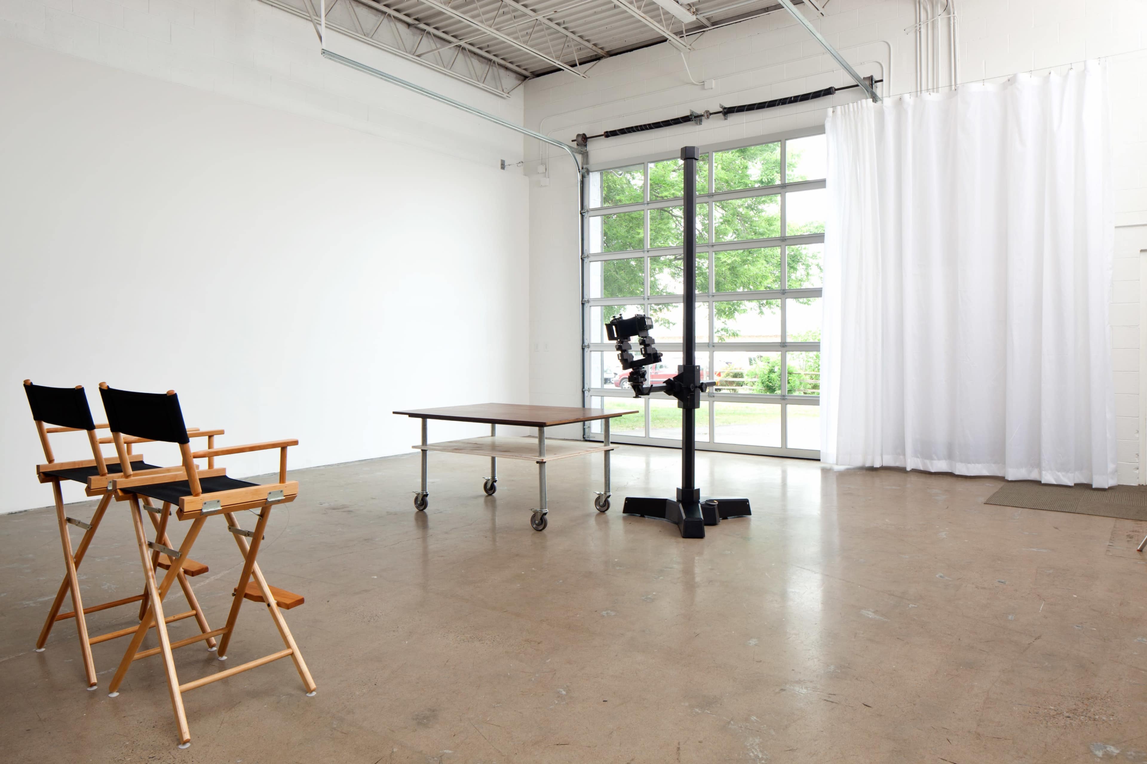 The image shows a minimalist studio space featuring two director's chairs, a table on wheels, and a camera setup in front of a large window with curtains.