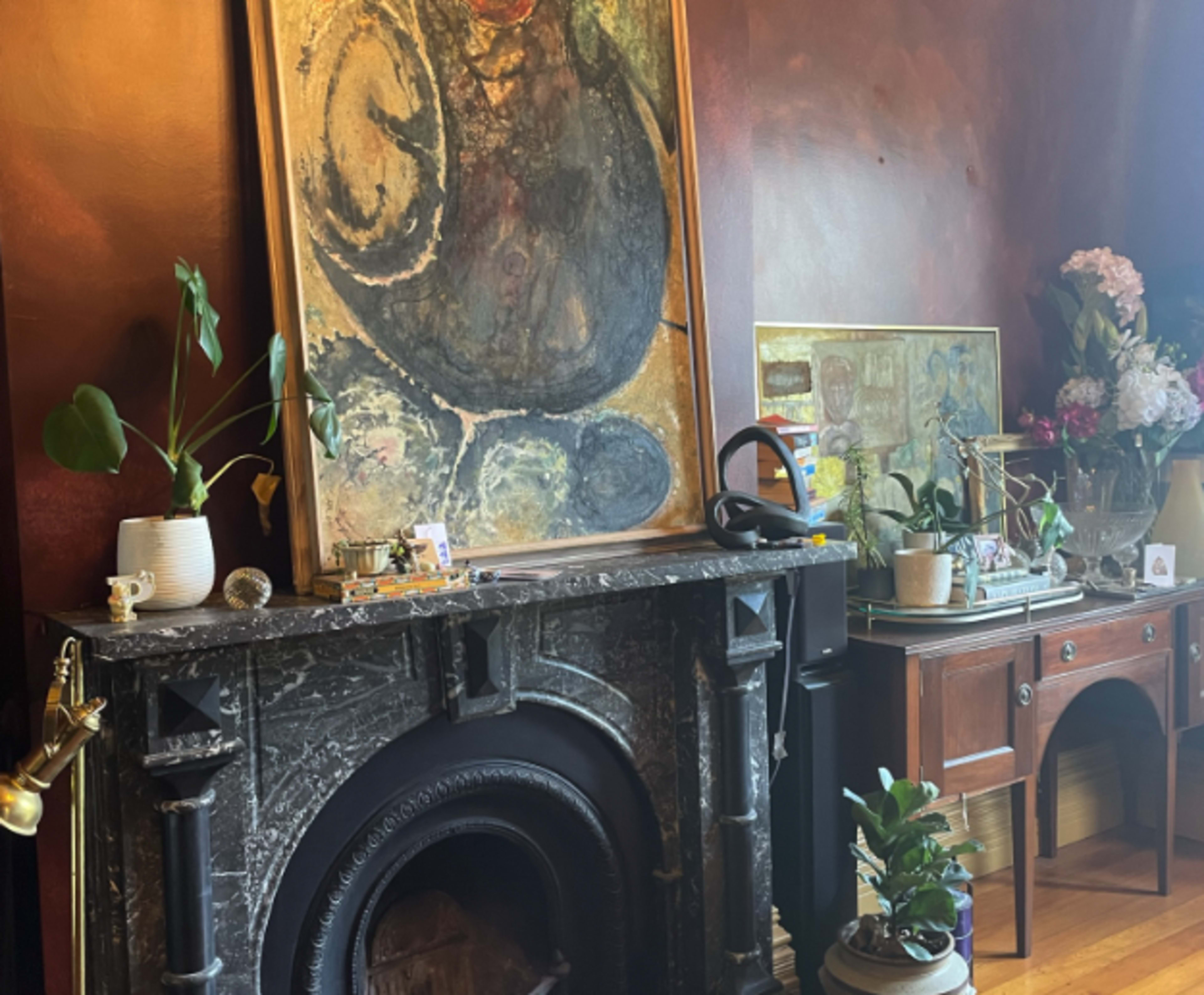 A dark-hued room with a large abstract painting above a marble fireplace, surrounded by various decorative items and plants.