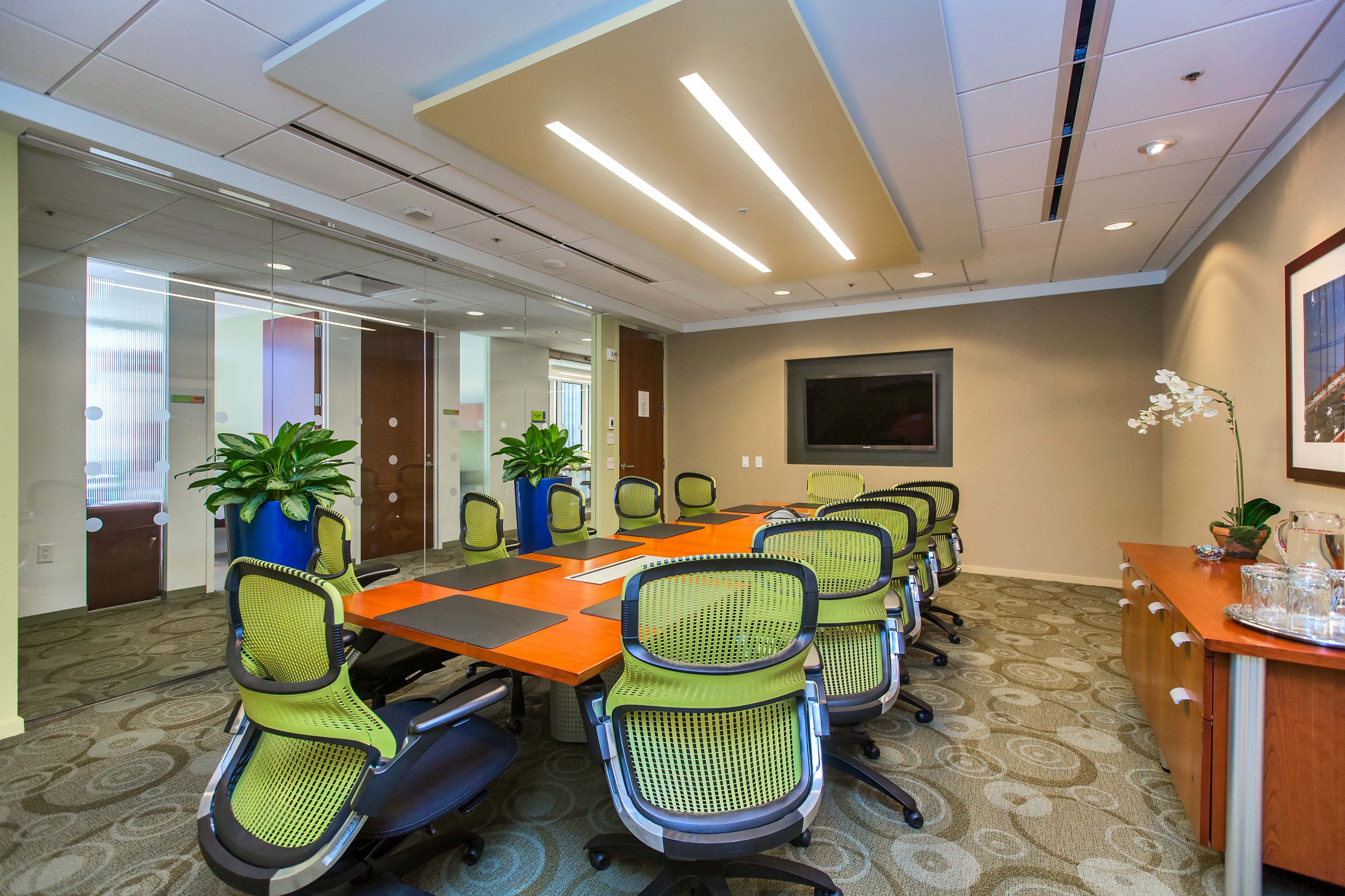A modern conference room with a large orange table surrounded by green office chairs and plants, featuring glass walls and a mounted screen.