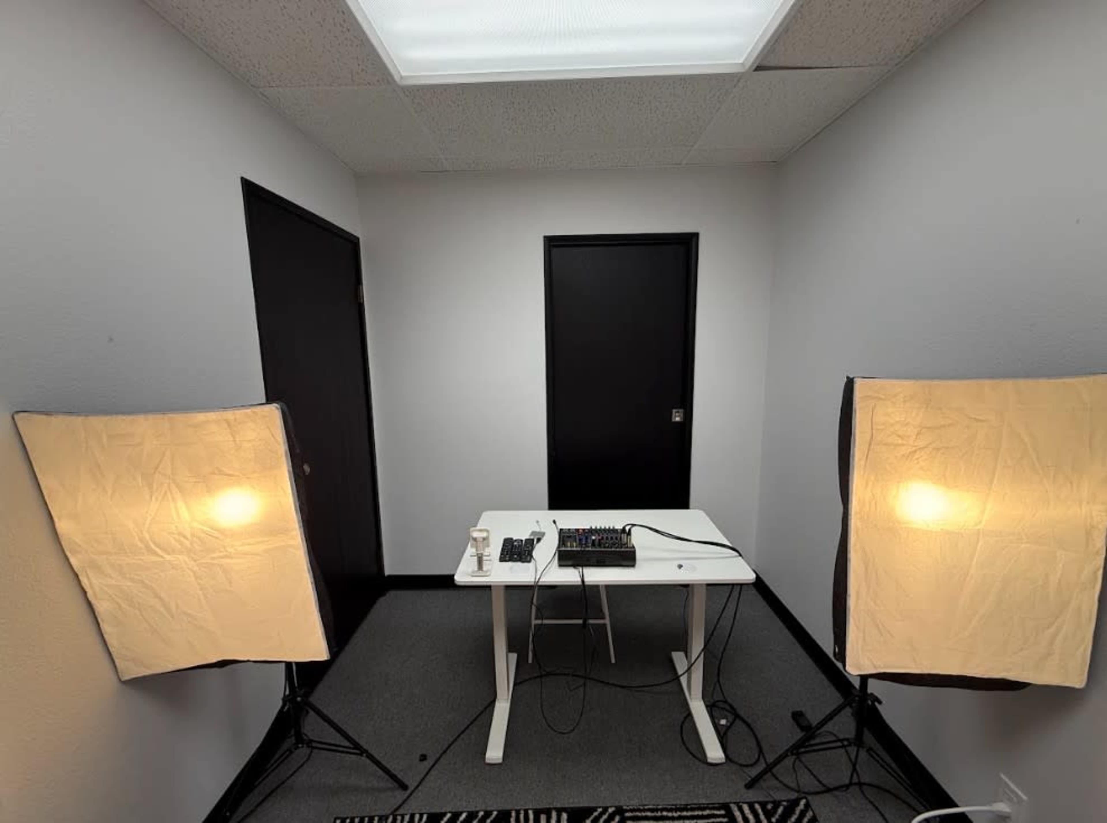 A small, sparsely furnished room features a white table with audio equipment and two softbox lights positioned on either side.