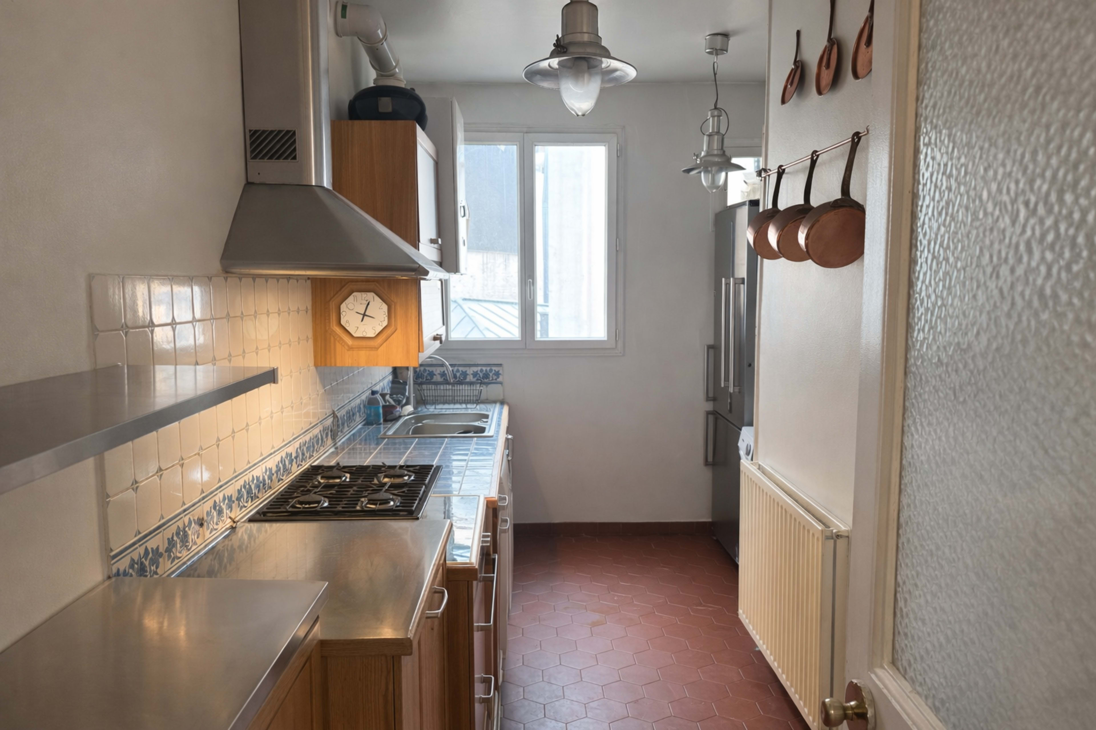 The image shows a compact kitchen with wooden cabinets, a stainless steel countertop, a gas stove, and copper pots hanging on the wall.