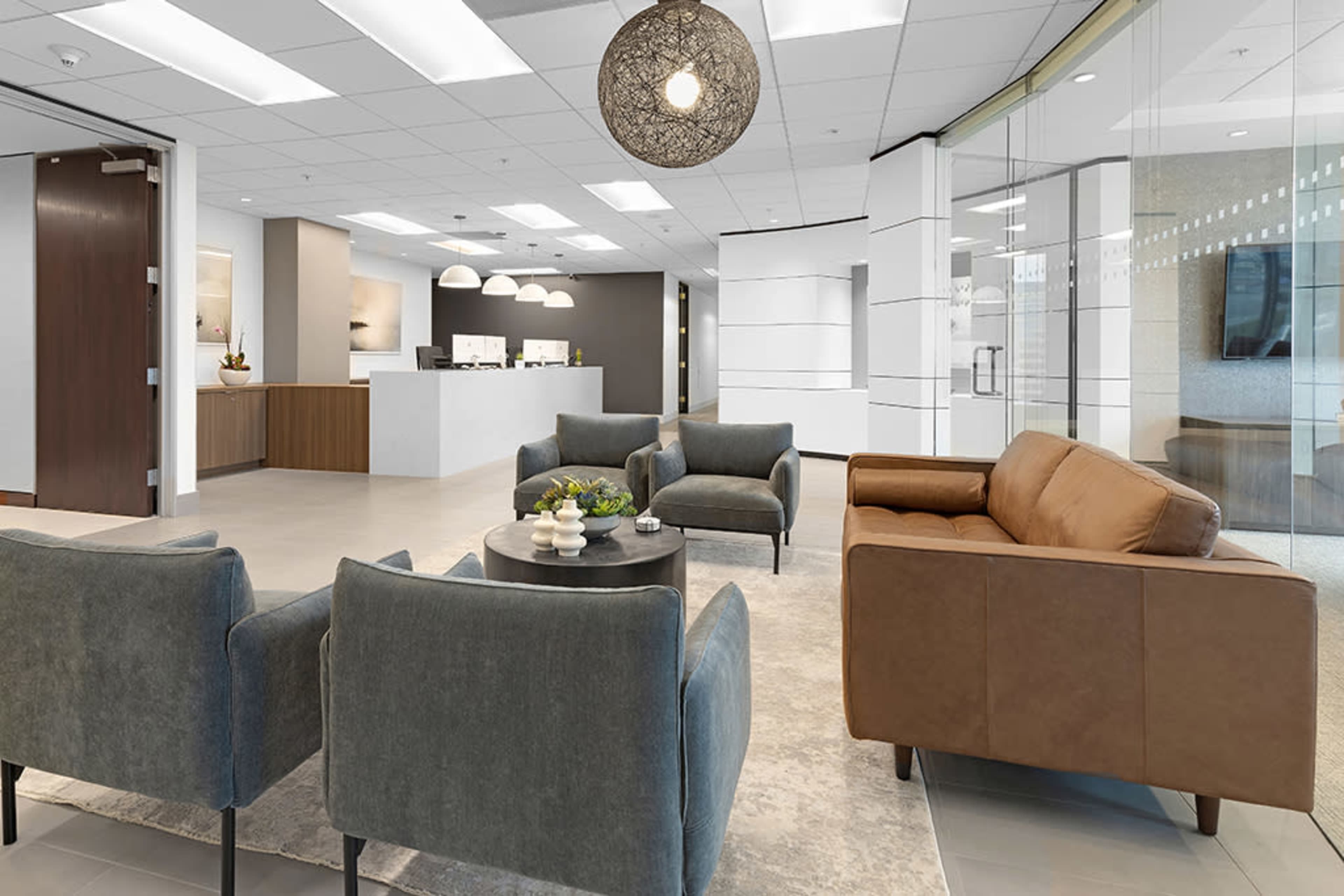 The image shows a modern office reception area with gray and brown seating, a circular coffee table, and a welcoming front desk in the background.