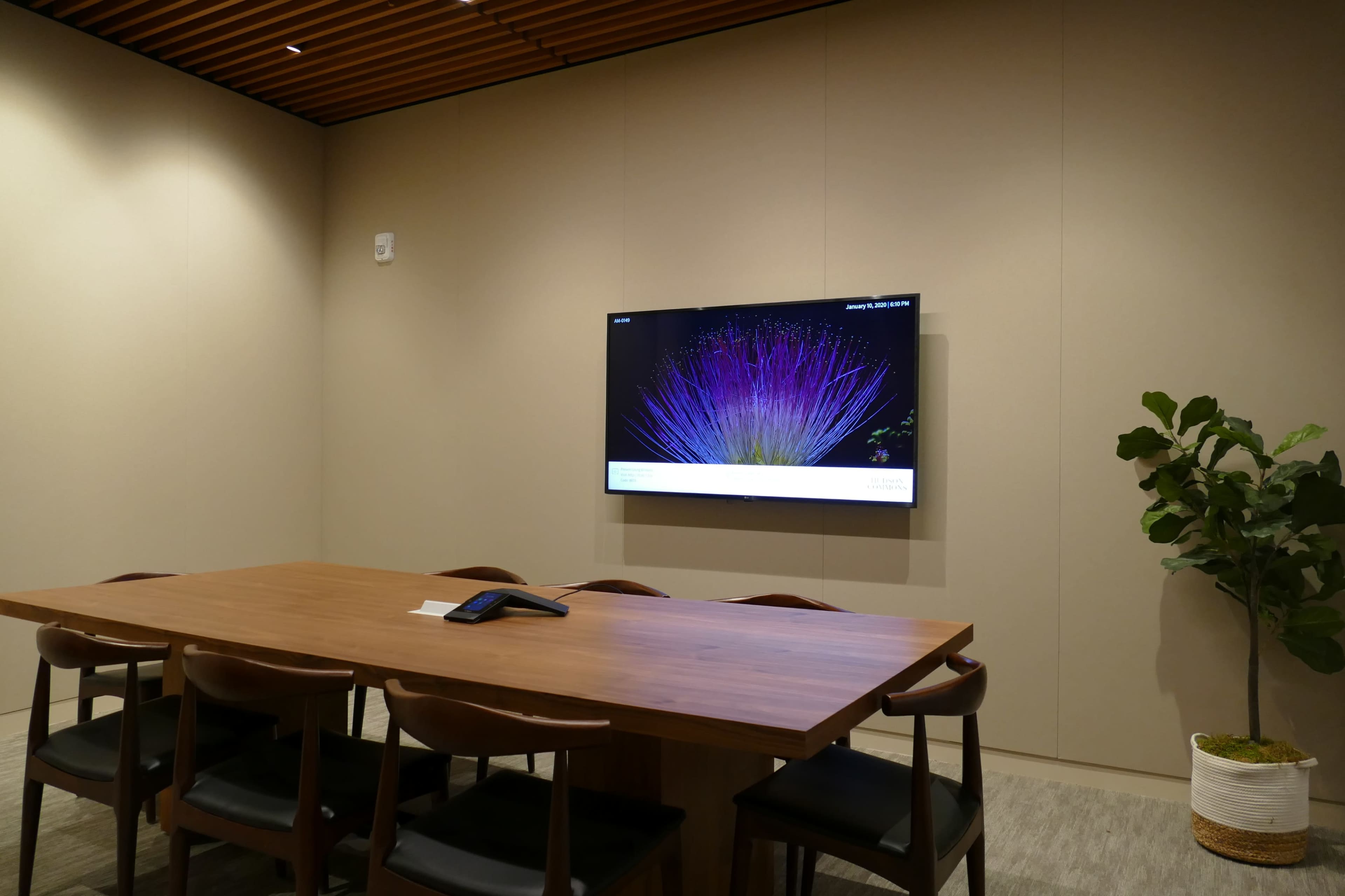 A conference room features a wooden table with six chairs, a wall-mounted screen displaying colorful graphics, and a potted plant in the corner.