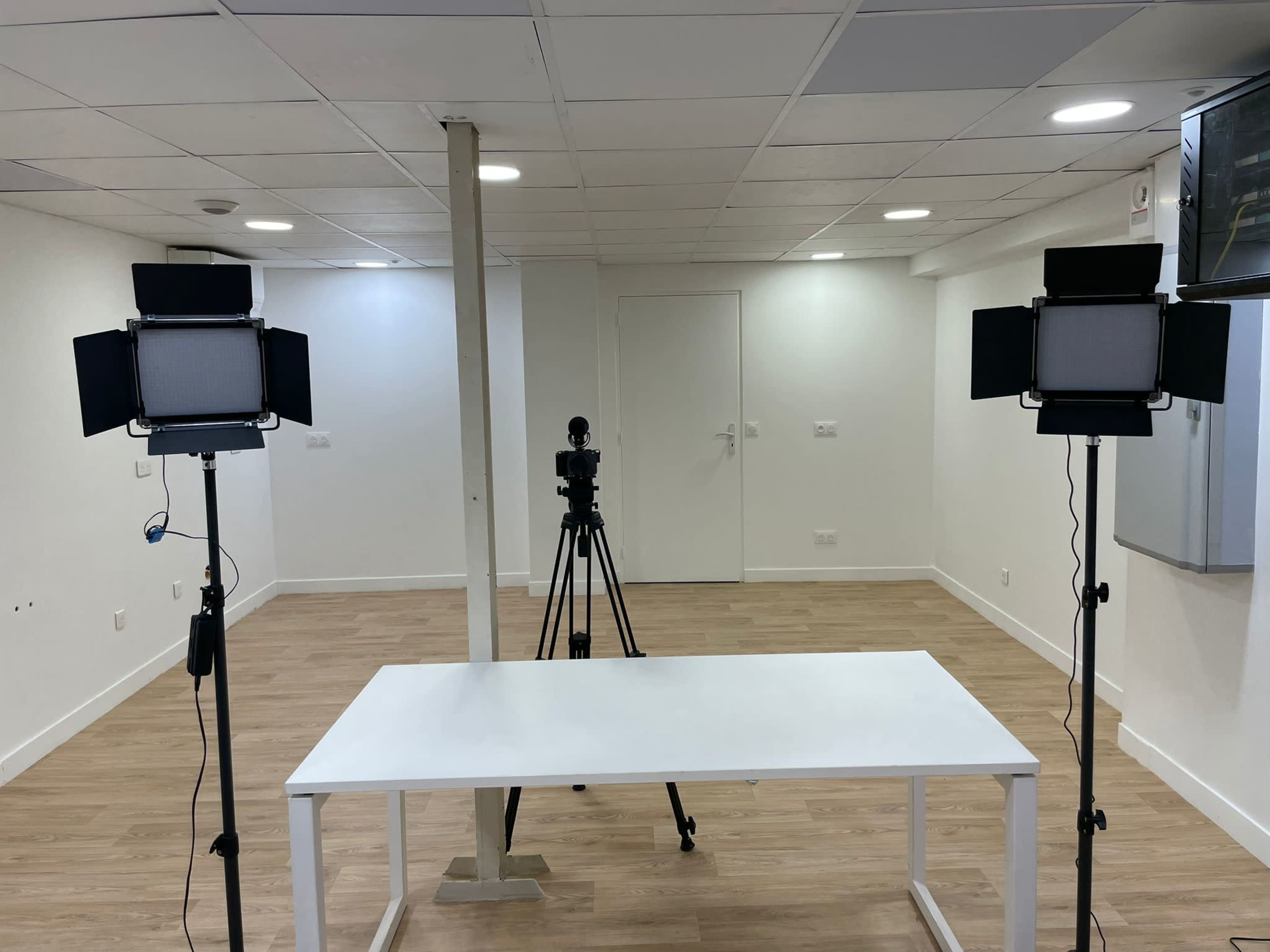 The image shows a well-lit, empty room with two studio lights positioned on either side of a white table, facing a camera on a tripod.