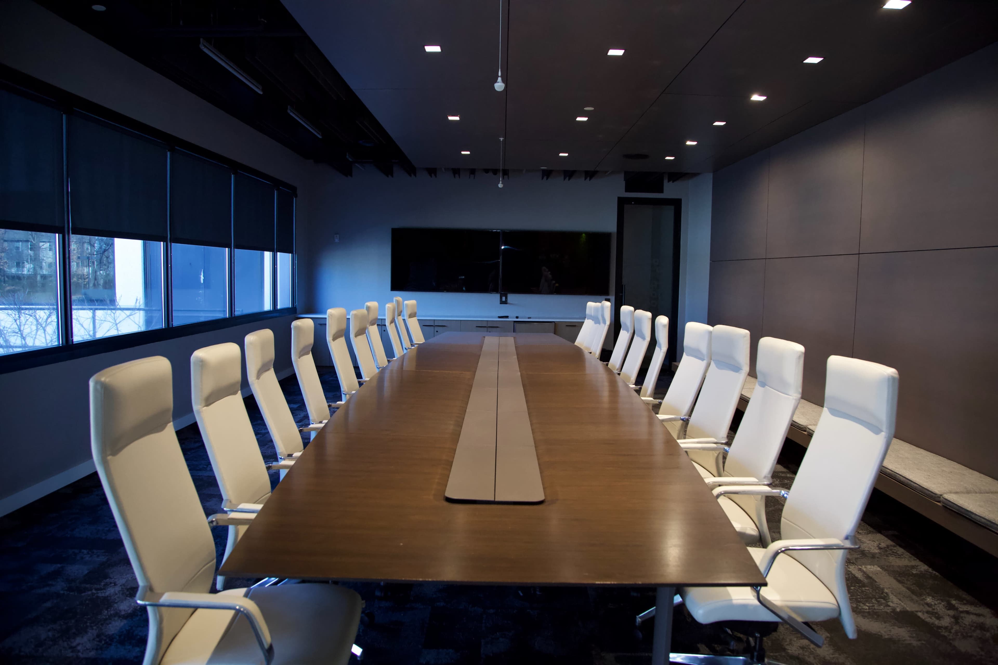 A long conference table with white chairs is set up in a modern meeting room featuring large windows and a wall-mounted screen.