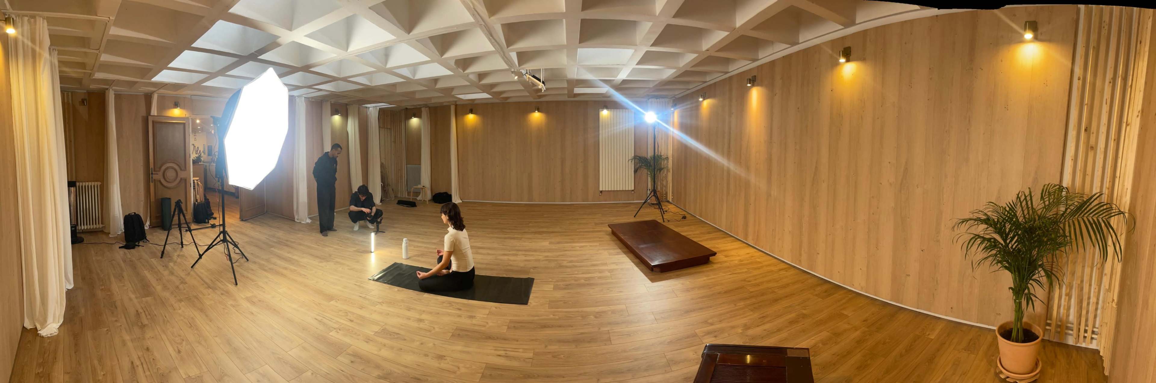 A spacious, well-lit studio with wooden walls and flooring features a woman seated on a yoga mat, while a photographer and an assistant prepare for a shoot.