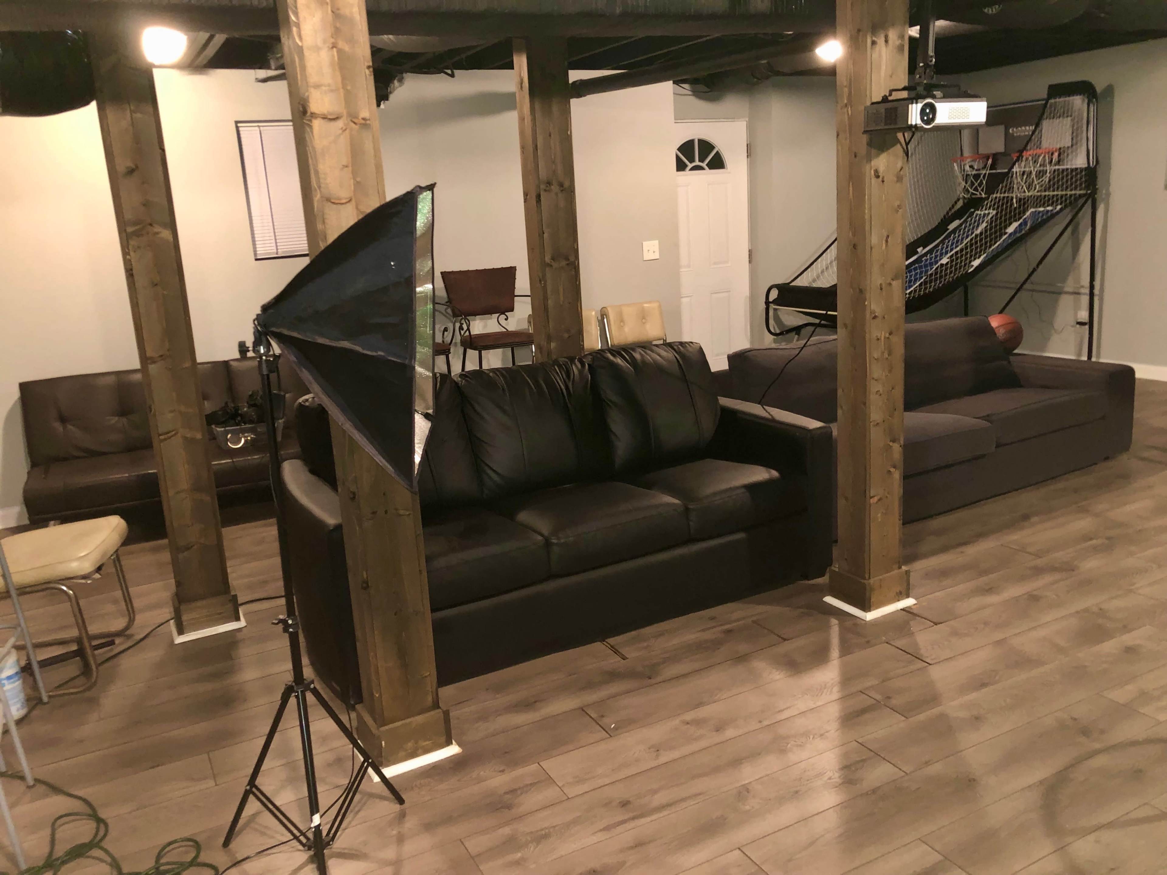 A spacious basement living area with two couches, wooden support beams, and a projector setup.