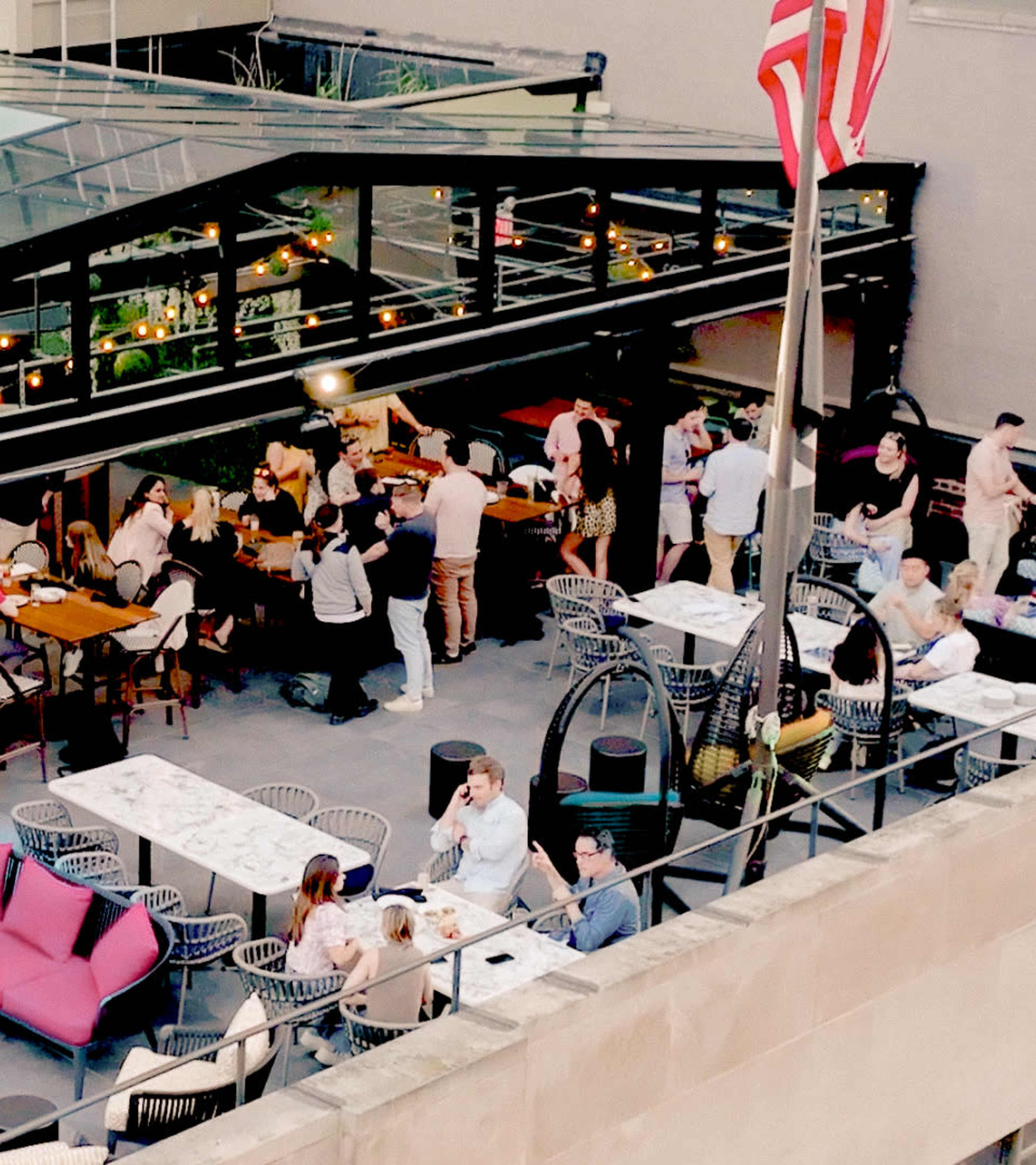 A rooftop bar scene features groups of people socializing around tables, with string lights illuminating the area and an American flag in the foreground.