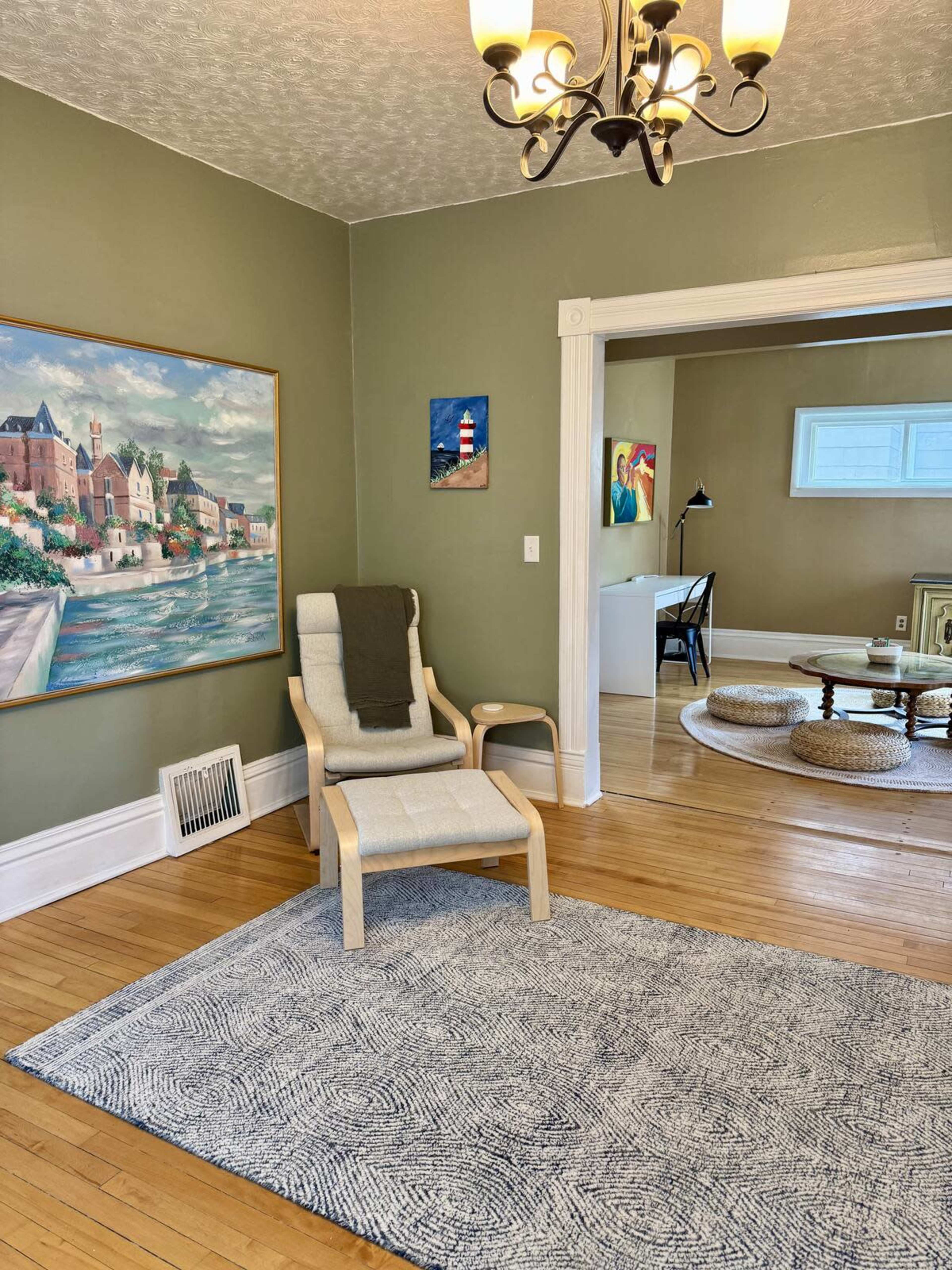 The image shows a living room with a large painting on the wall, a light-colored armchair, and hardwood floors, with a doorway leading to another room featuring a table and artworks.
