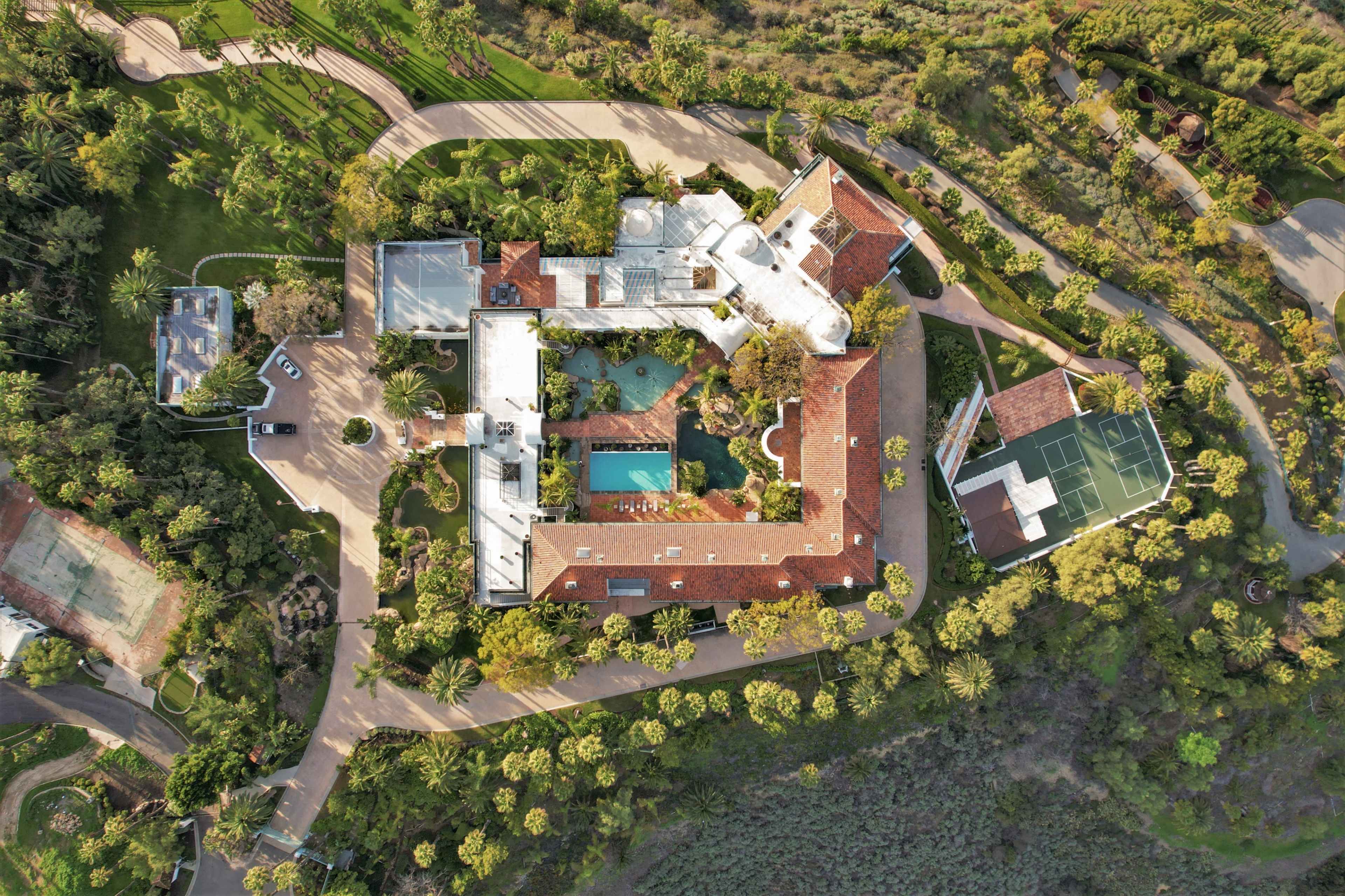 An aerial view of a large property featuring a main house, a swimming pool, tennis court, and landscaped gardens surrounded by palm trees.