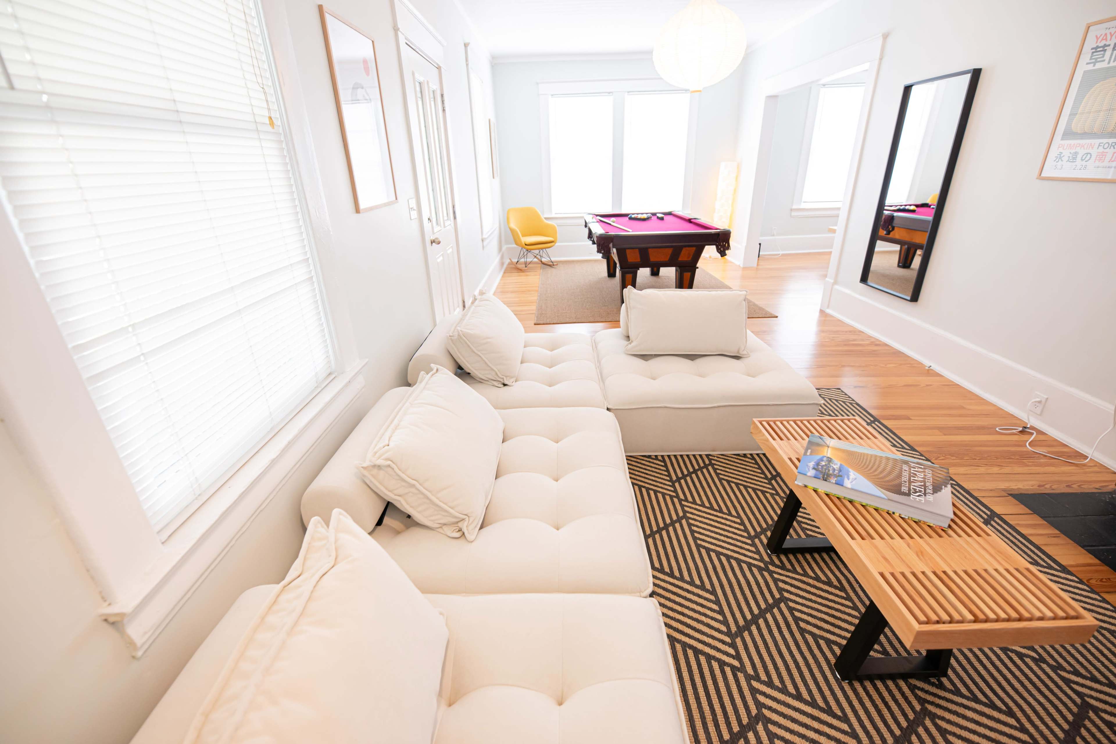 A modern living room features a white sectional sofa, a wooden coffee table, and a pool table in the background.