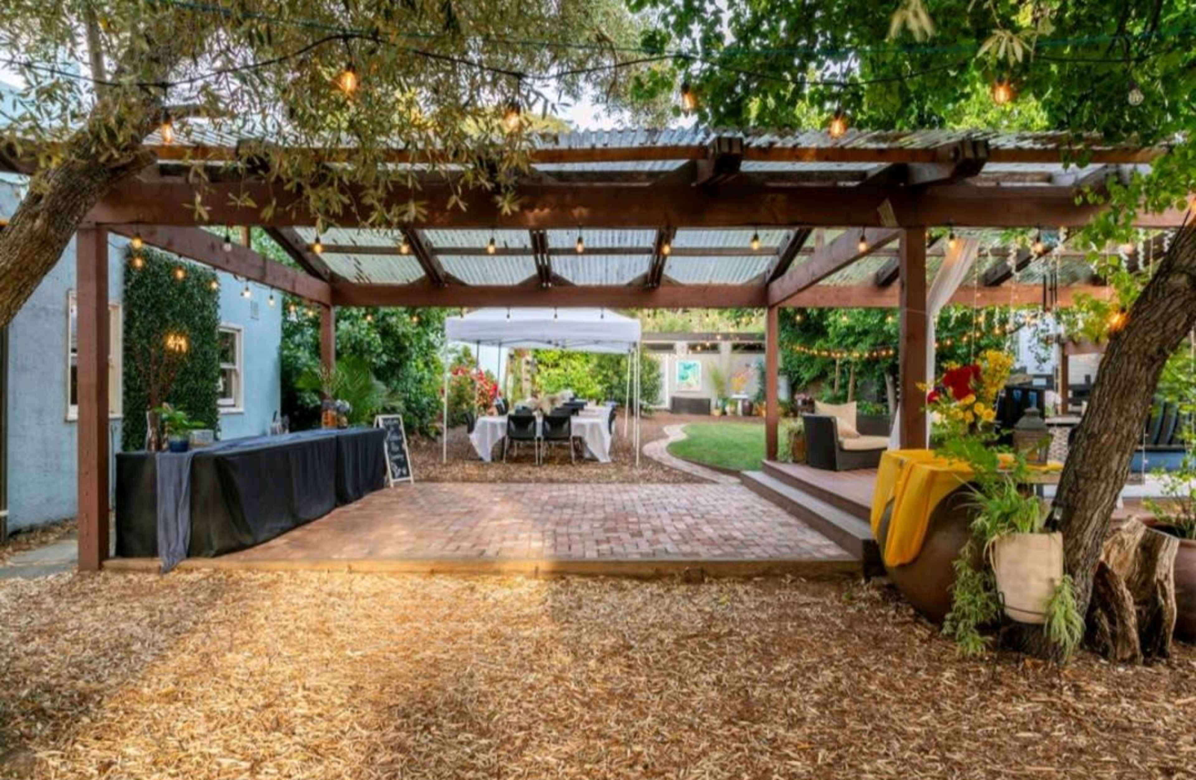 A spacious outdoor patio features a wooden pergola with string lights, surrounded by greenery and furnished with tables and seating areas.