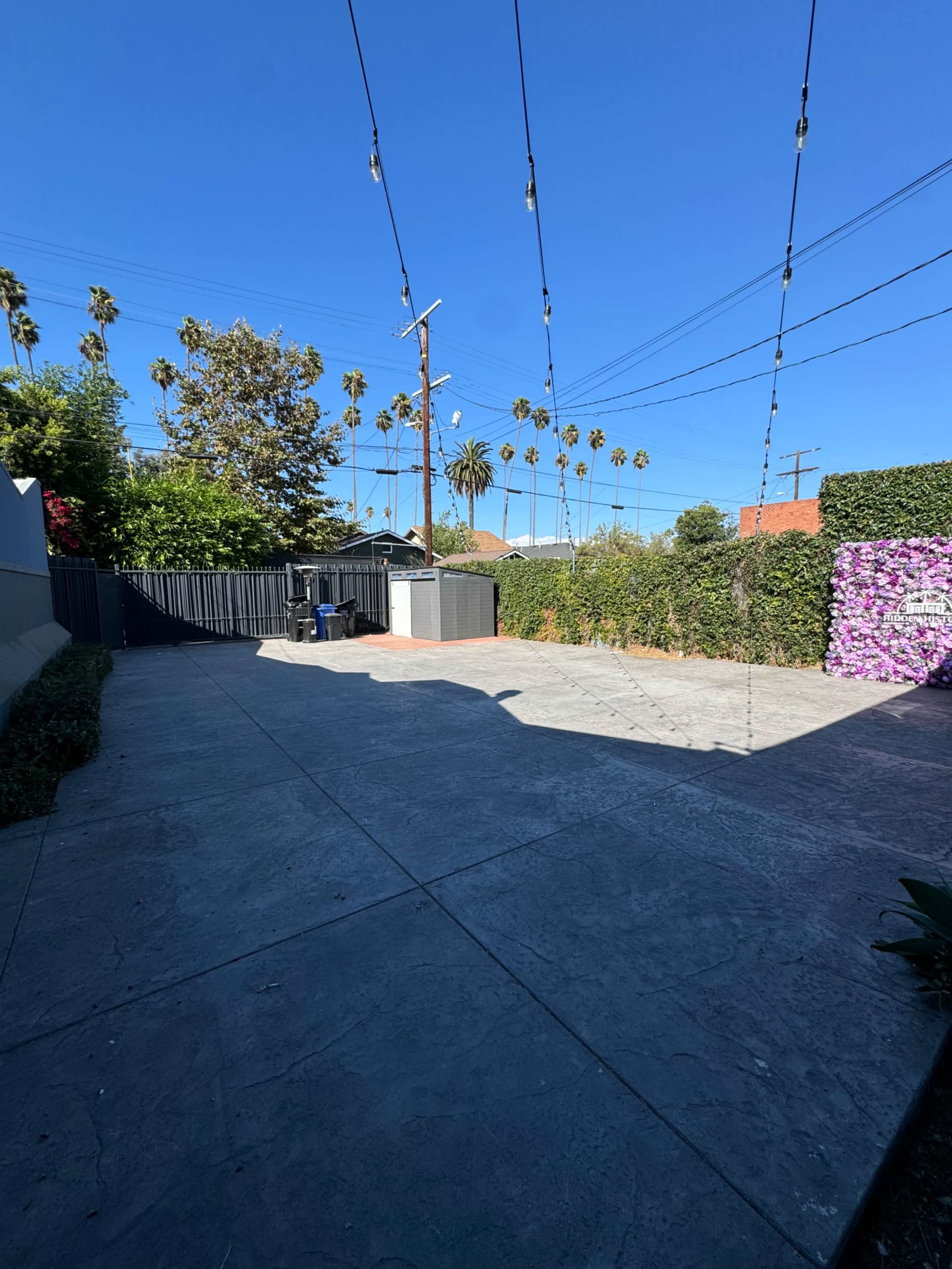 The image shows a spacious, empty backyard with concrete flooring, surrounded by greenery and palm trees under a clear blue sky.