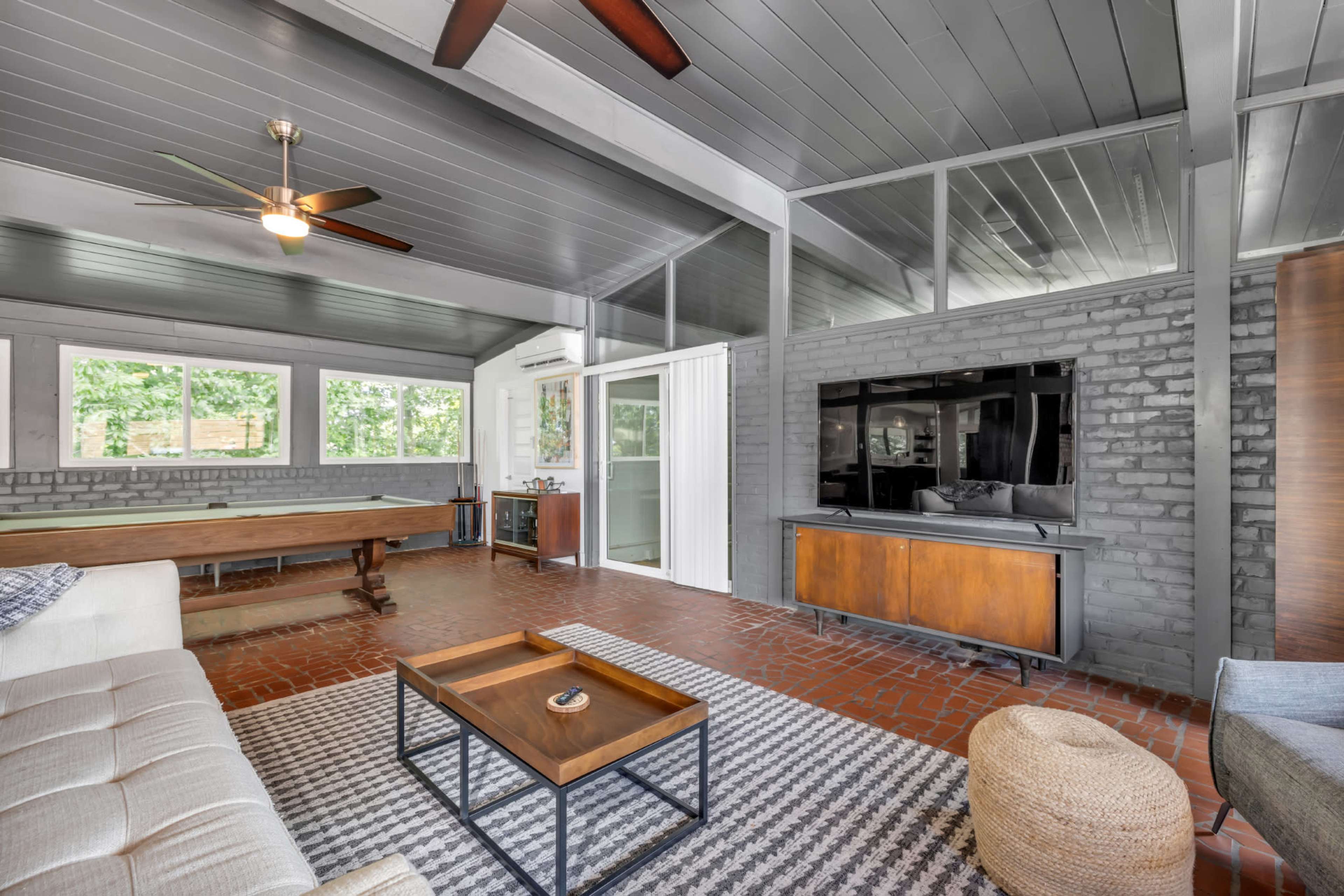 A modern living room with a brick accent wall, a flat-screen TV on a wooden console, a large window providing natural light, and a pool table in the background.