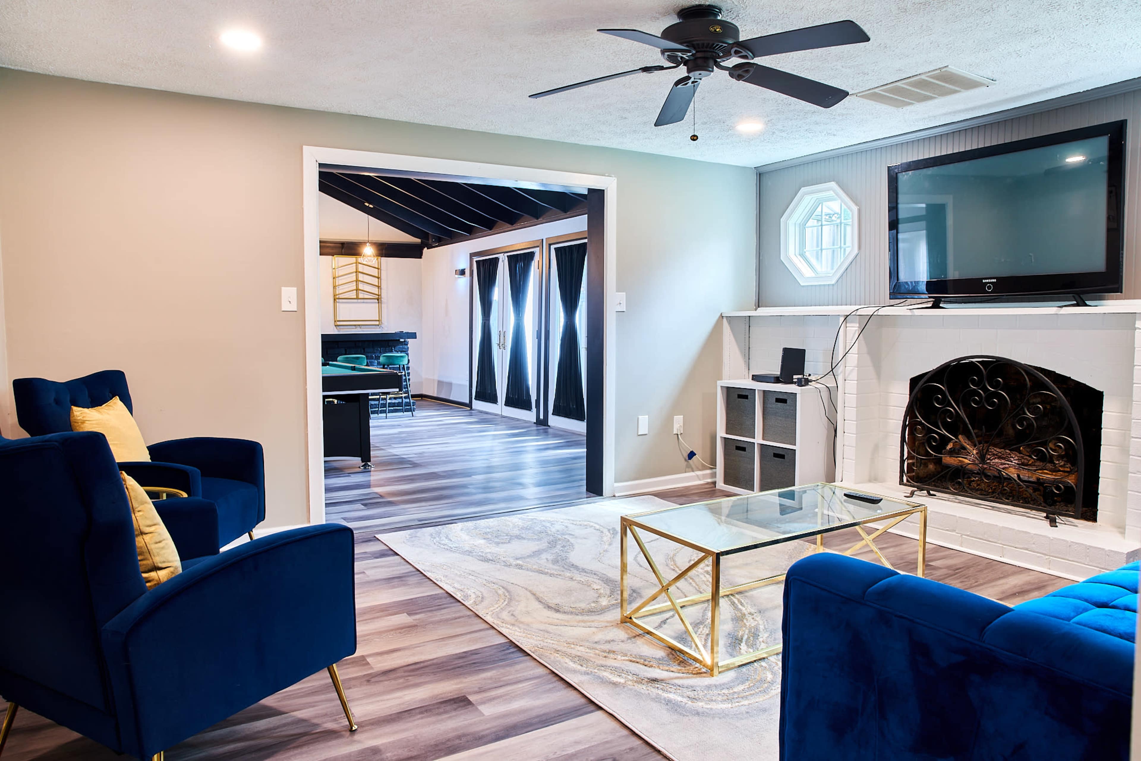 A modern living room with blue armchairs, a glass coffee table, a television mounted on the wall, and a view of an adjoining room through an open doorway.