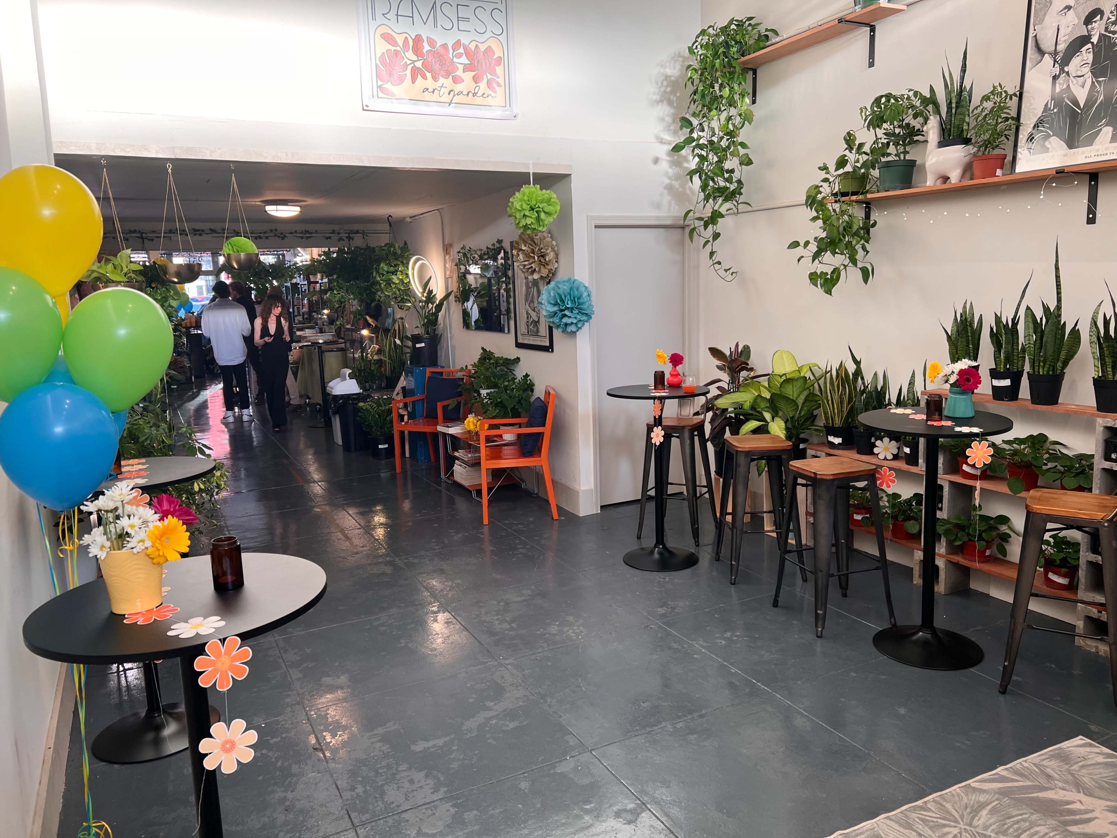 A brightly decorated indoor space with potted plants, tables adorned with flower decorations, and colorful balloons.