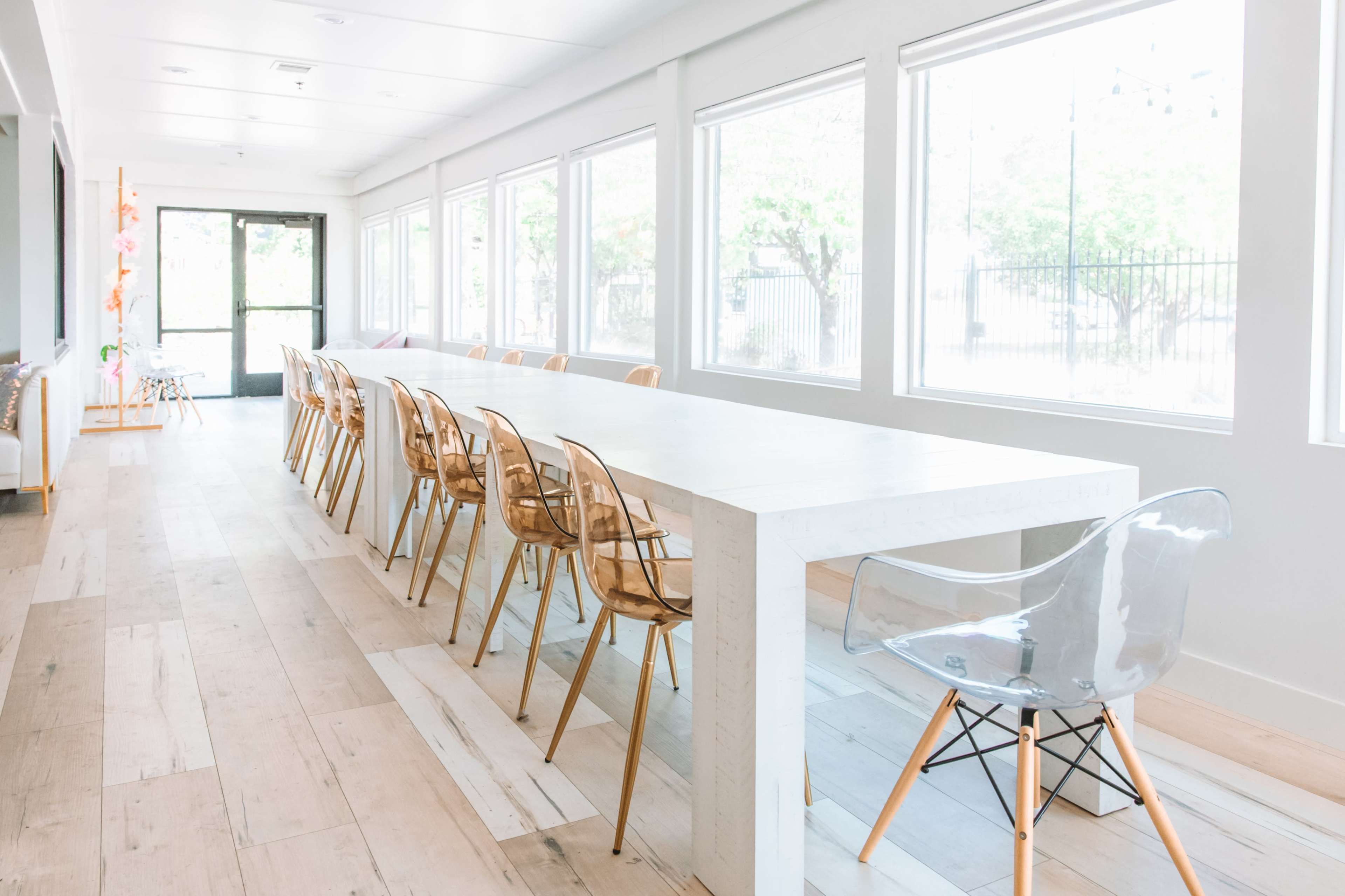 A long, white table with transparent chairs is arranged in a brightly lit room with large windows.