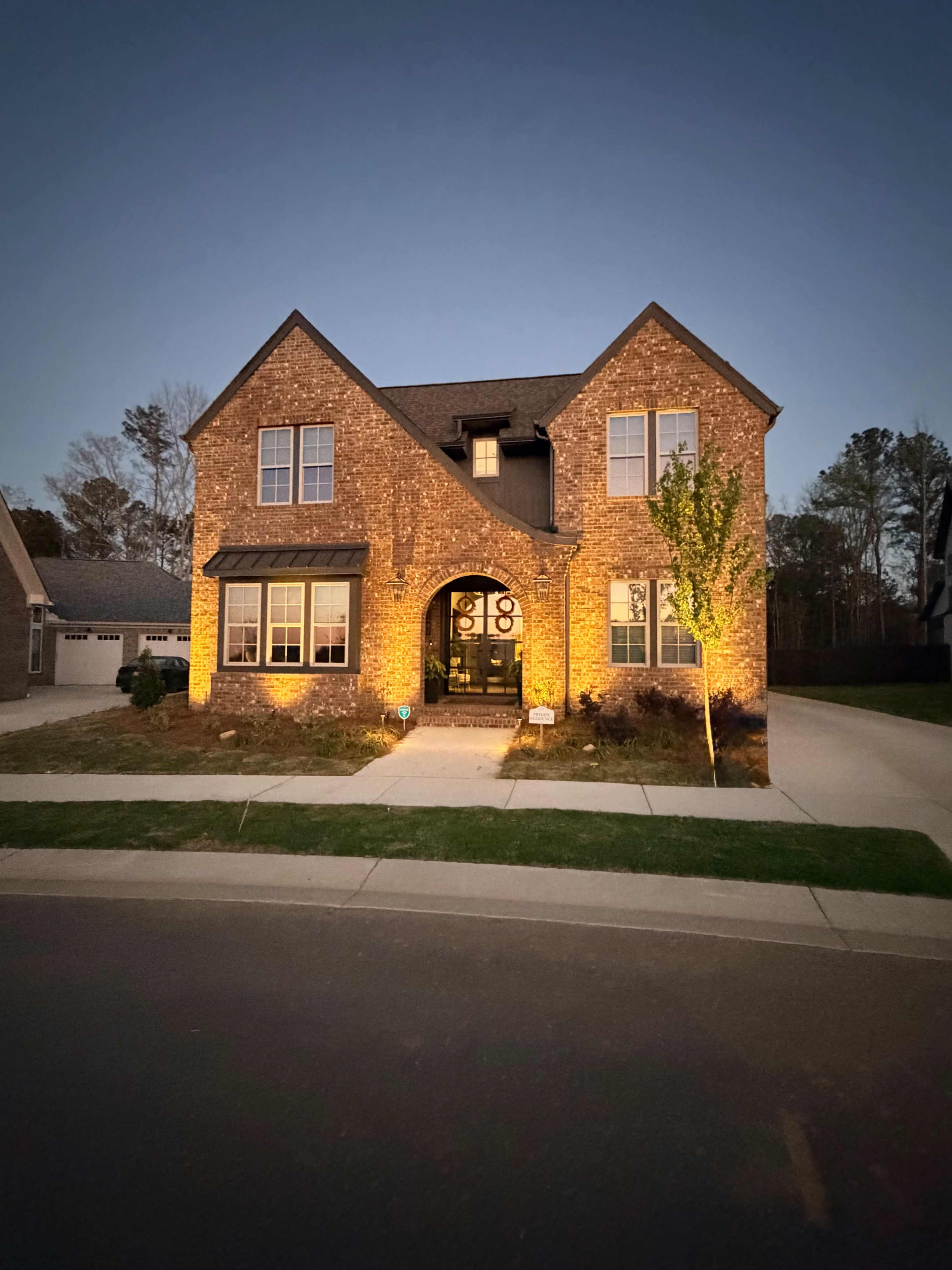 A two-story brick house is illuminated by exterior lighting as dusk falls, with a well-maintained lawn and driveway visible.