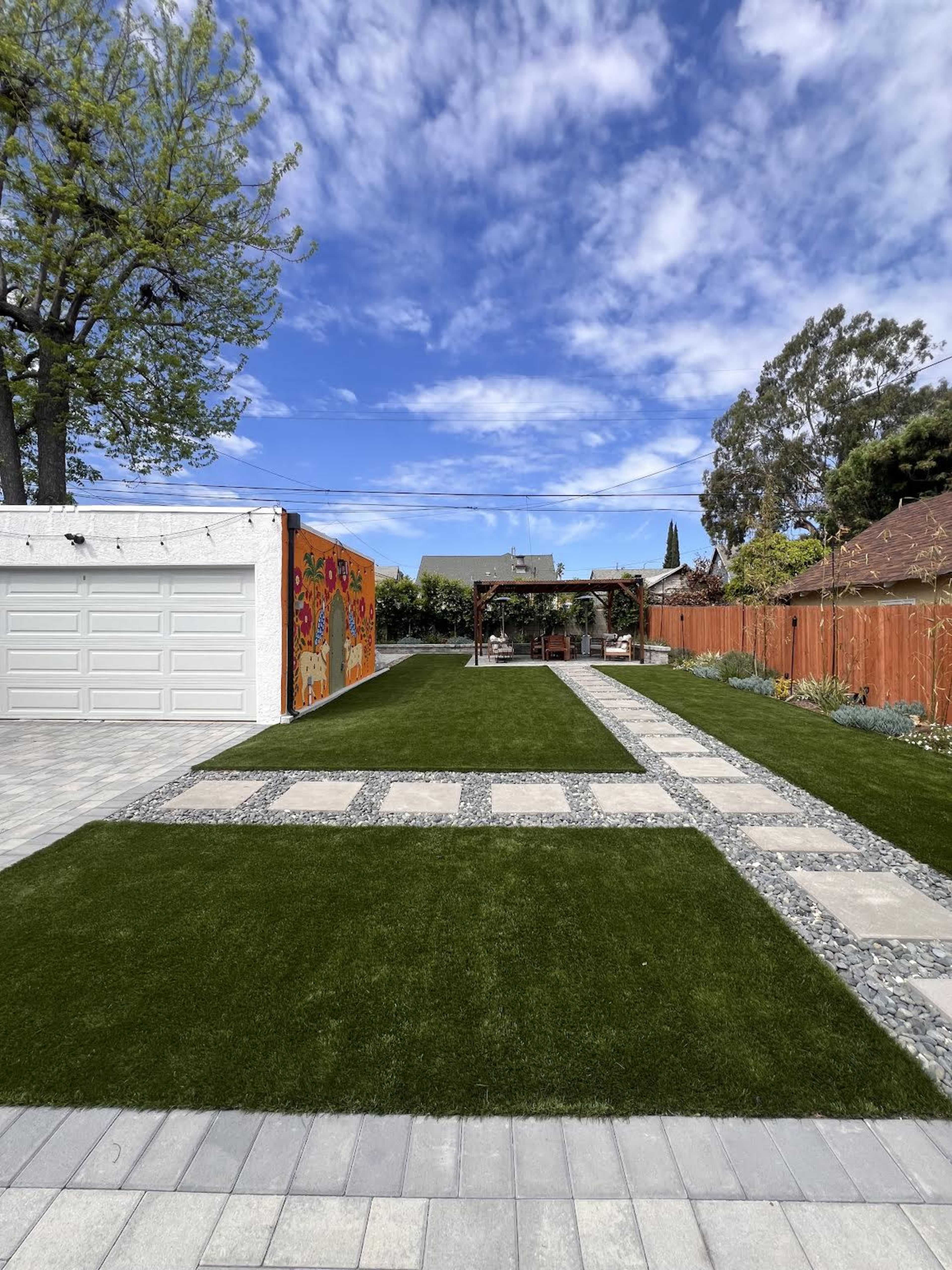 The image shows a landscaped backyard with artificial grass, a stone pathway, and a wooden pergola at the far end, surrounded by trees and a colorful mural on one side.