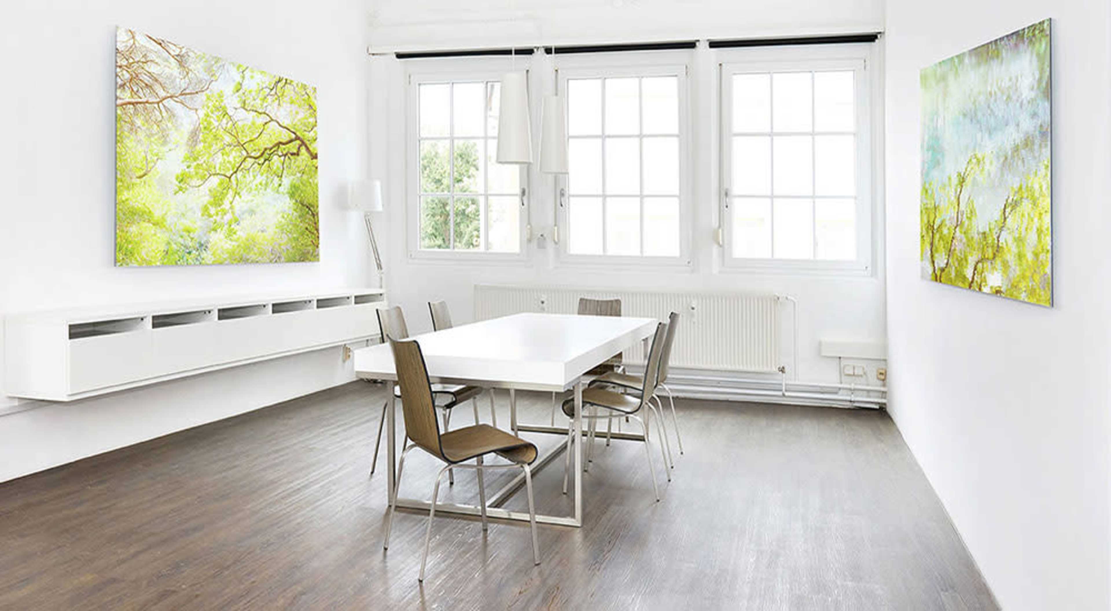 The image shows a bright, minimalist conference room featuring a white table surrounded by metal chairs, with large artworks hanging on the walls.