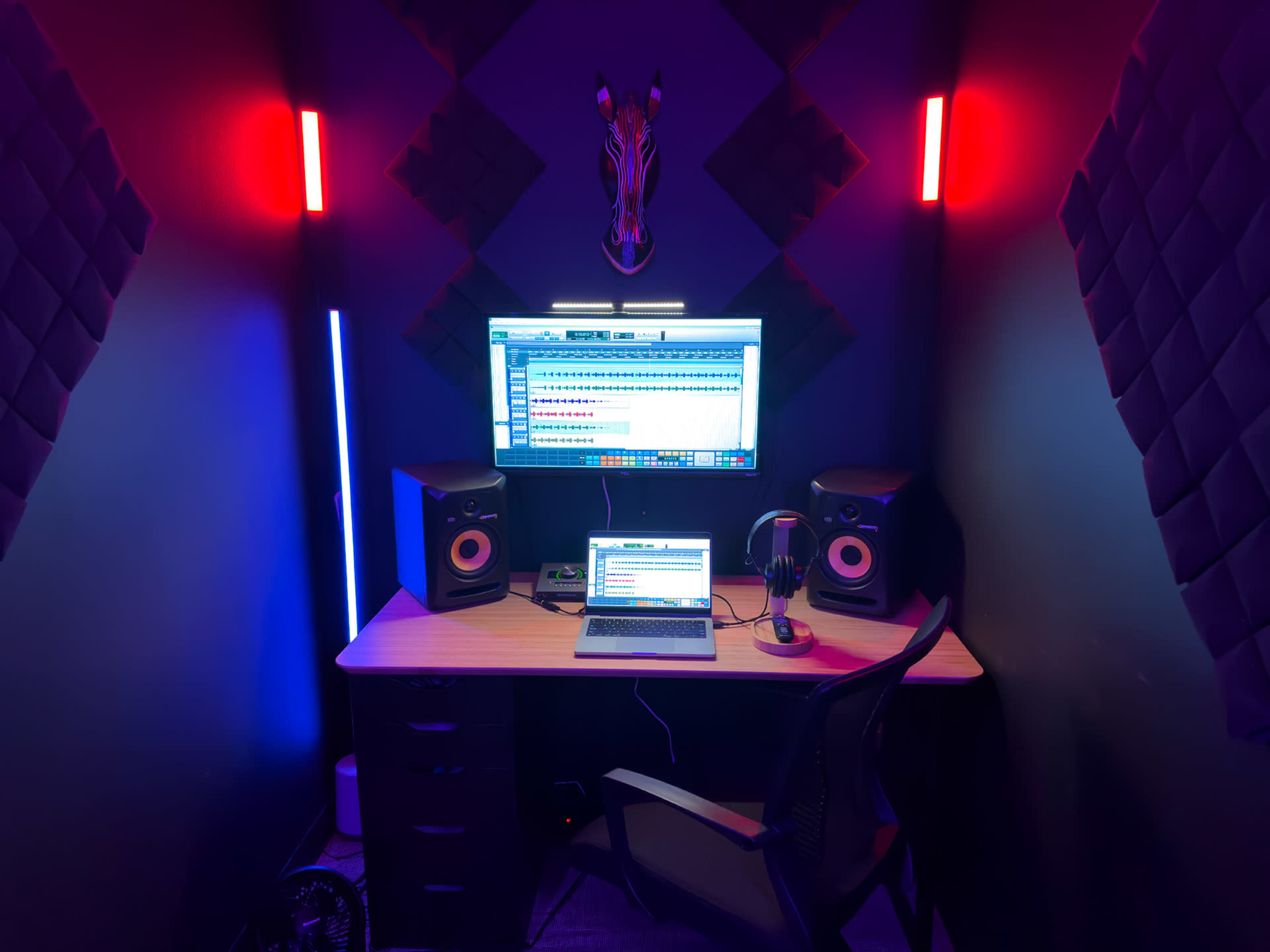 The image shows a small music production studio with a computer, speakers, and colorful LED lighting on the walls.