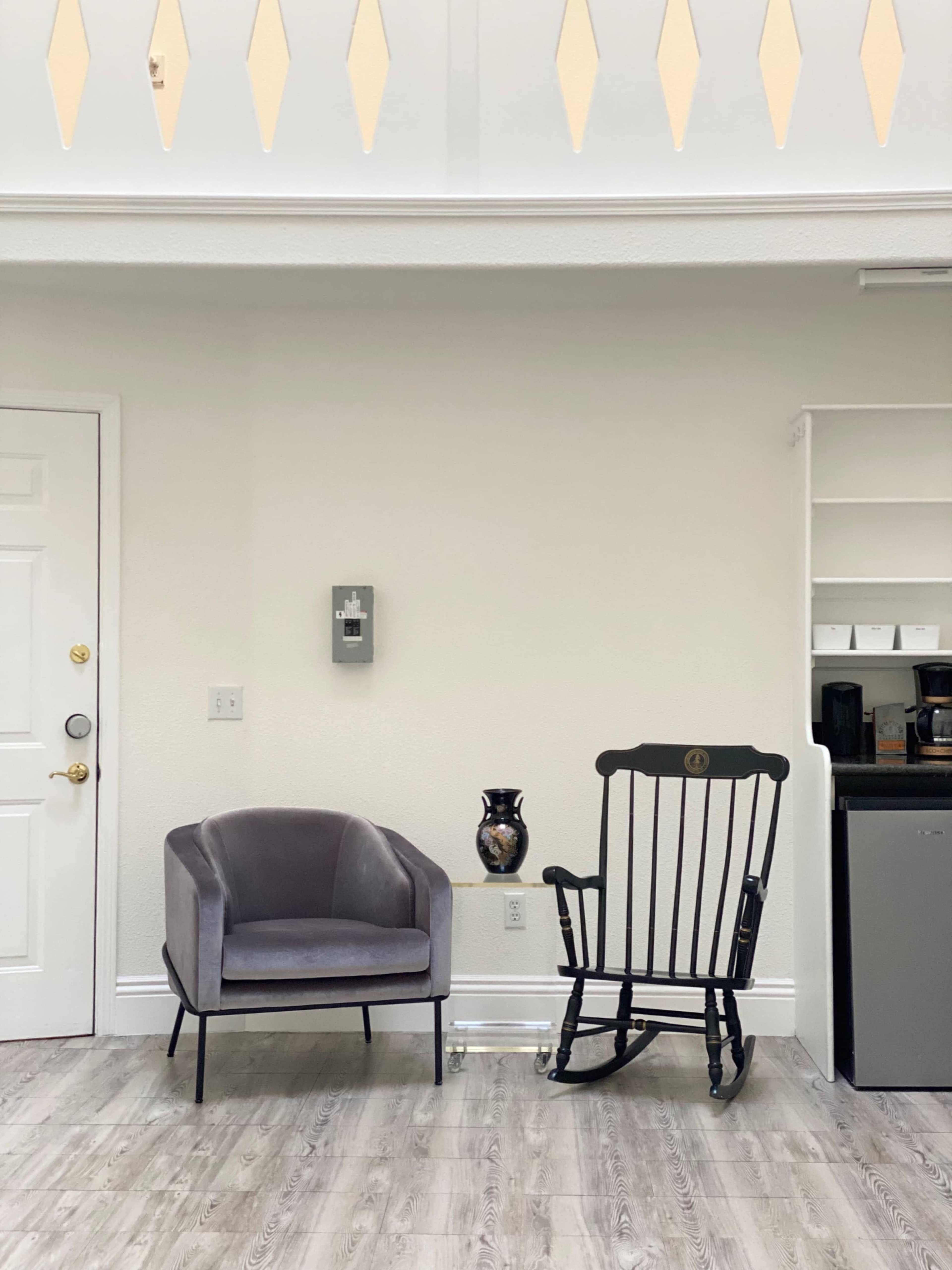 A gray armchair and a black rocking chair are placed against a light-colored wall in a room with a small kitchenette visible nearby.