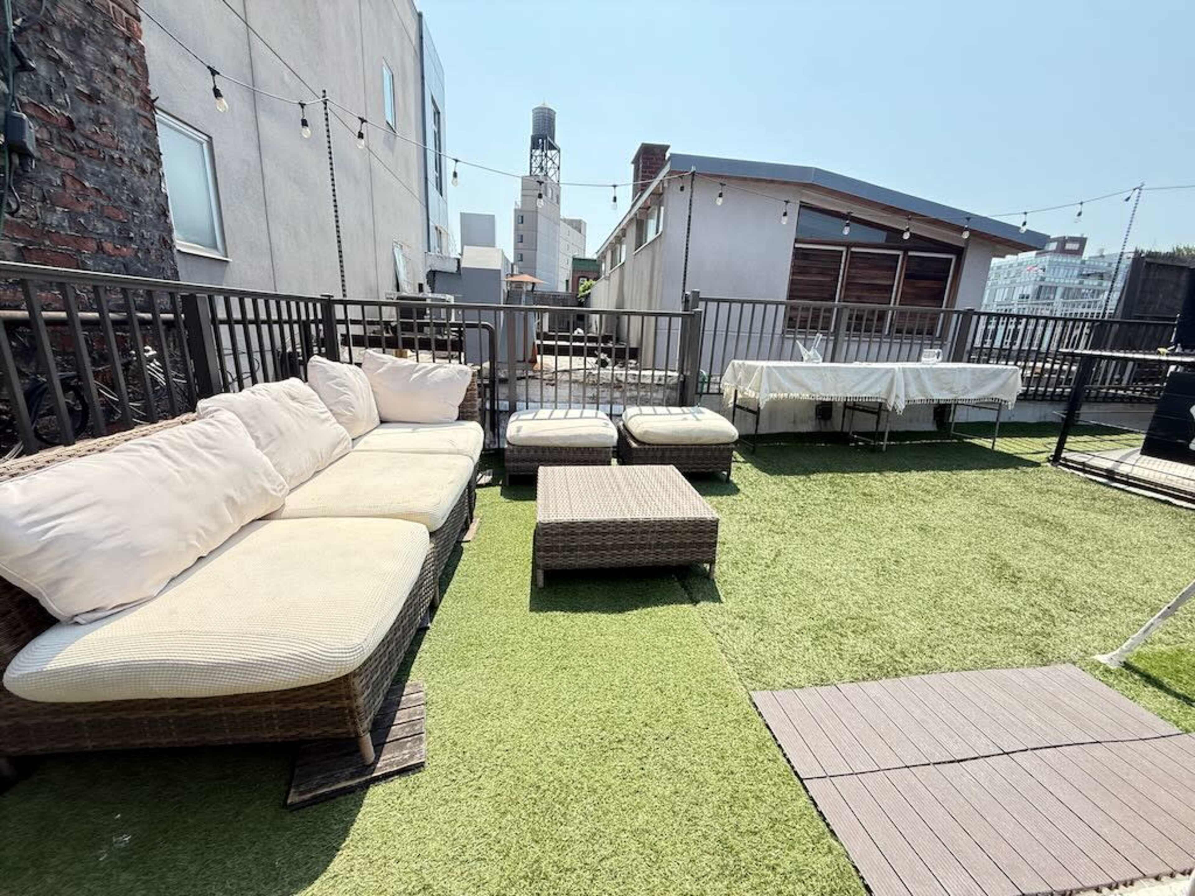 The image shows a rooftop patio area with a sectional sofa, a coffee table, and green artificial grass under a clear blue sky.