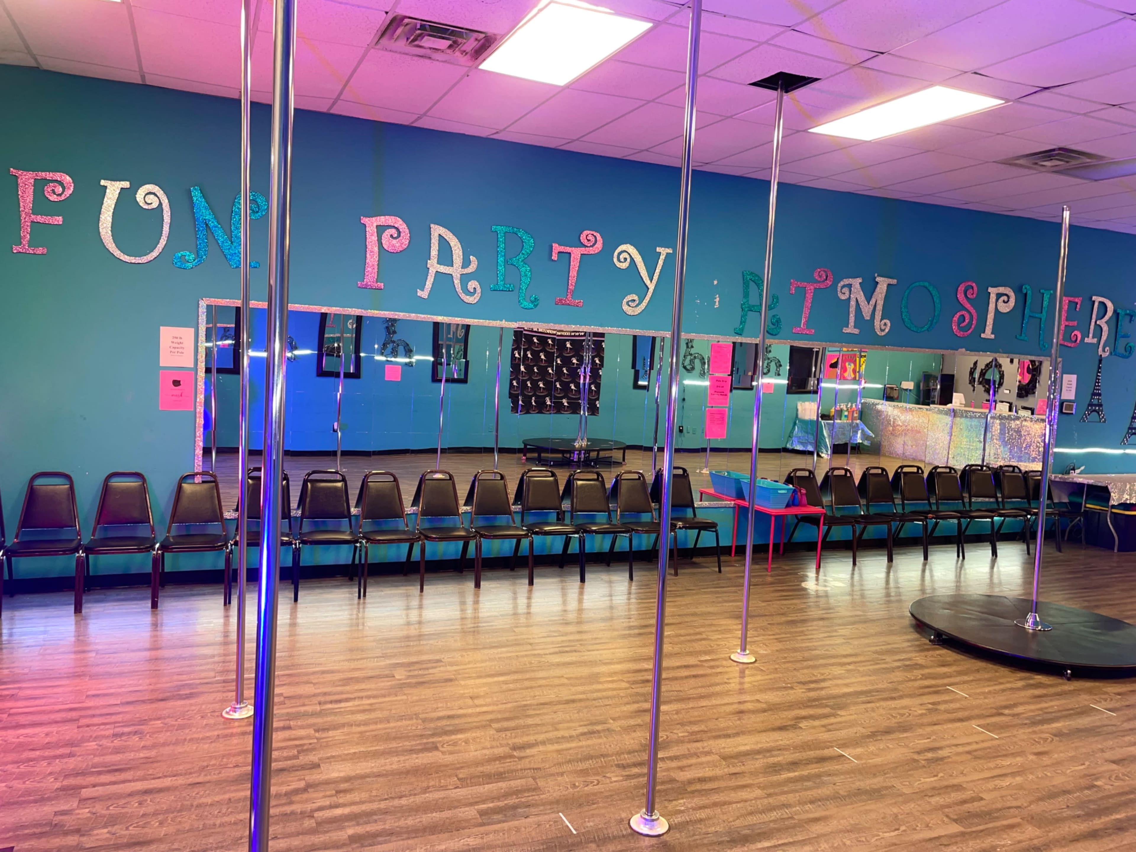 A brightly painted room with a mirrored wall, seating arranged in a row, and several poles for pole dancing.