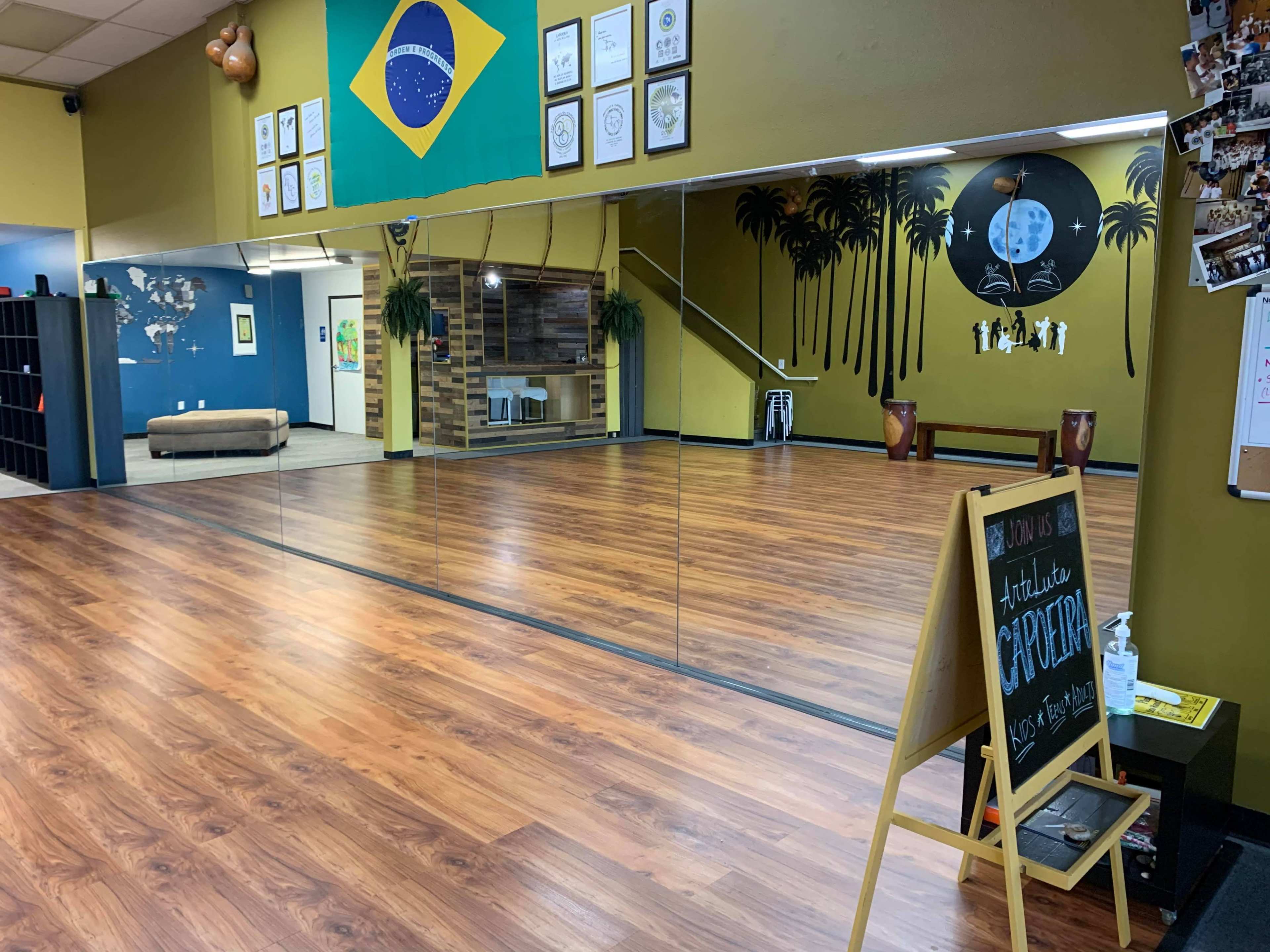 A spacious dance studio with wooden flooring, mirrors on one wall, a Brazilian flag hanging, and decorative elements like palm trees and a chalkboard sign.