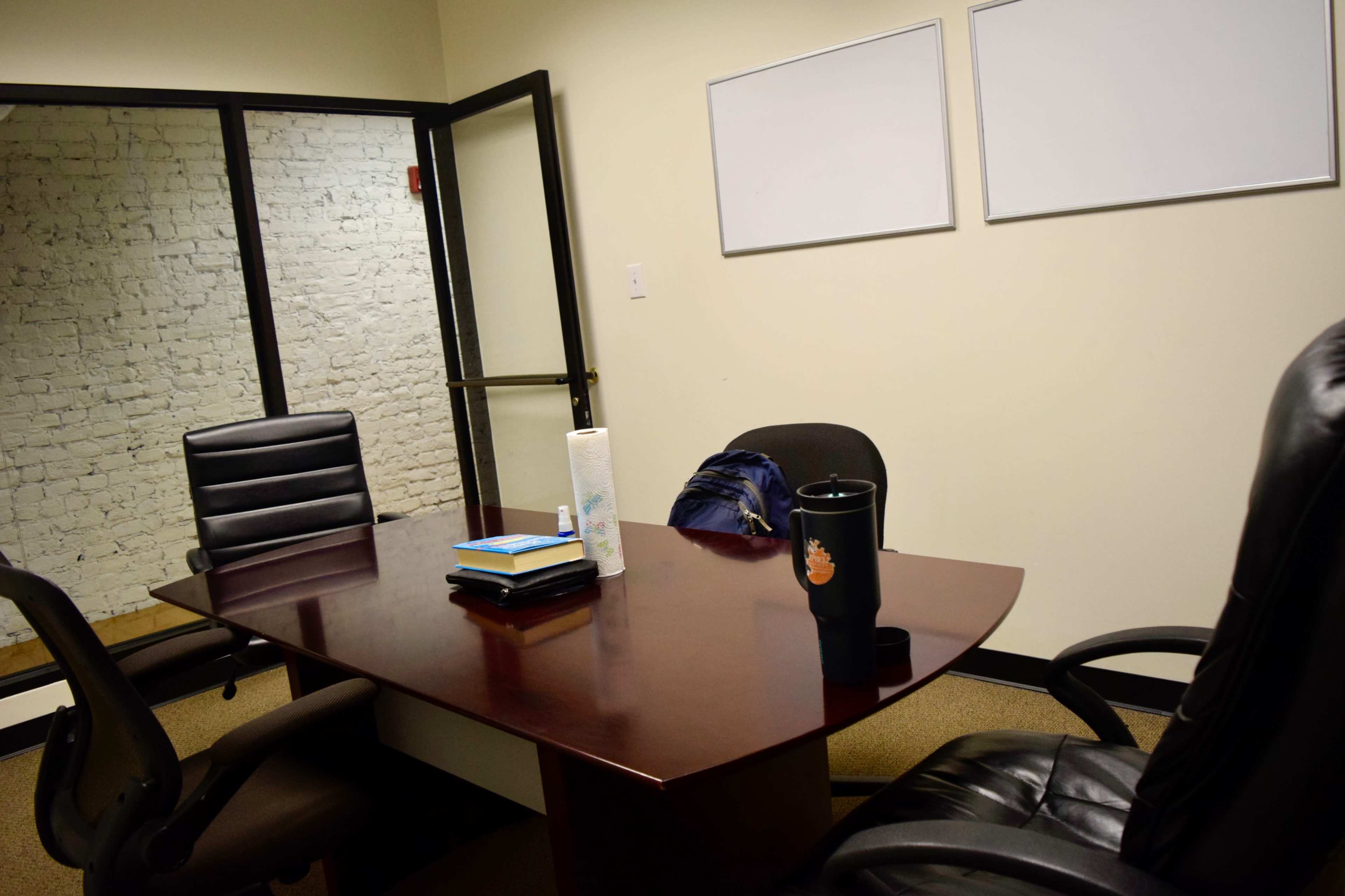 The image shows a small meeting room with a wooden table, three chairs, a stack of books, a roll of paper towels, and a drink container, with a glass wall reflecting a brick structure.