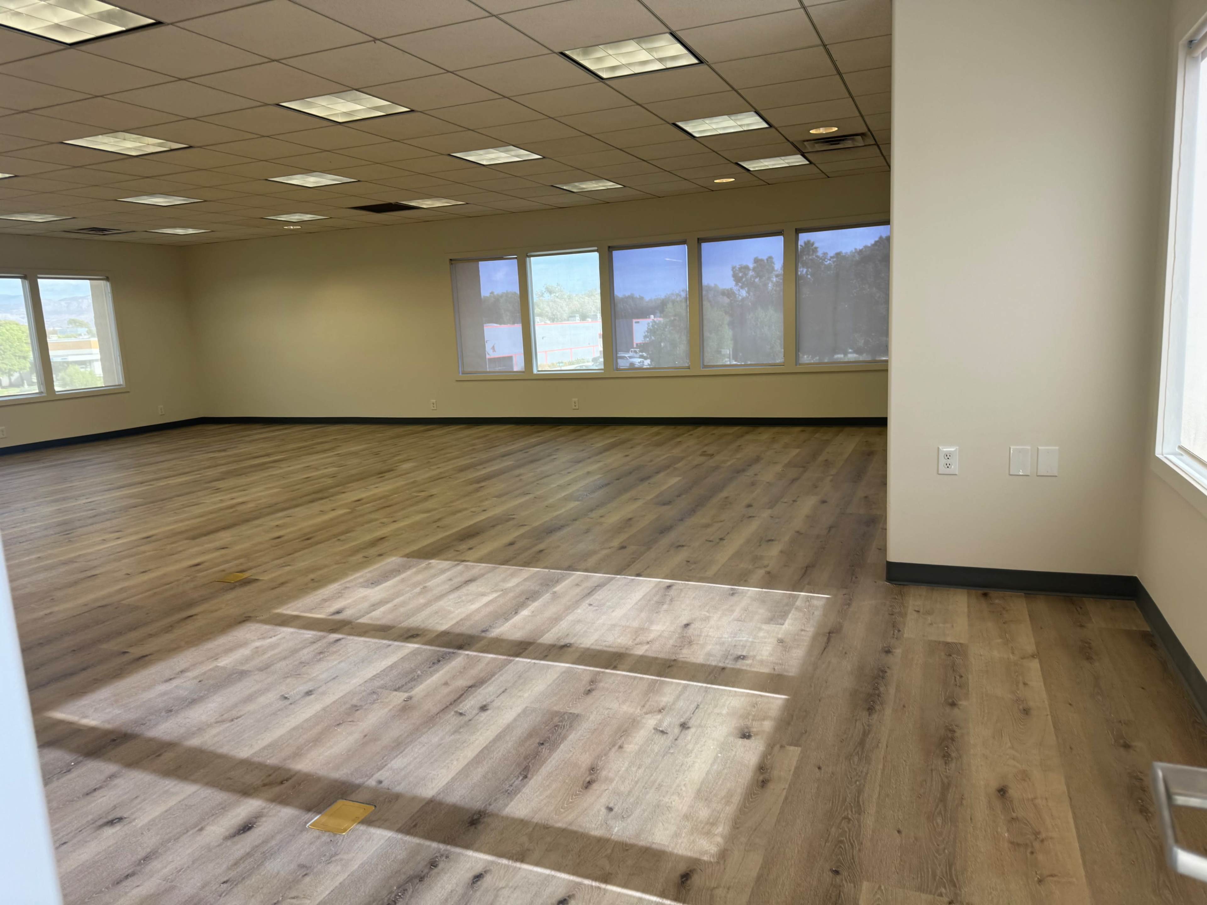 The image shows an empty office space with wooden flooring and large windows exposing natural light.