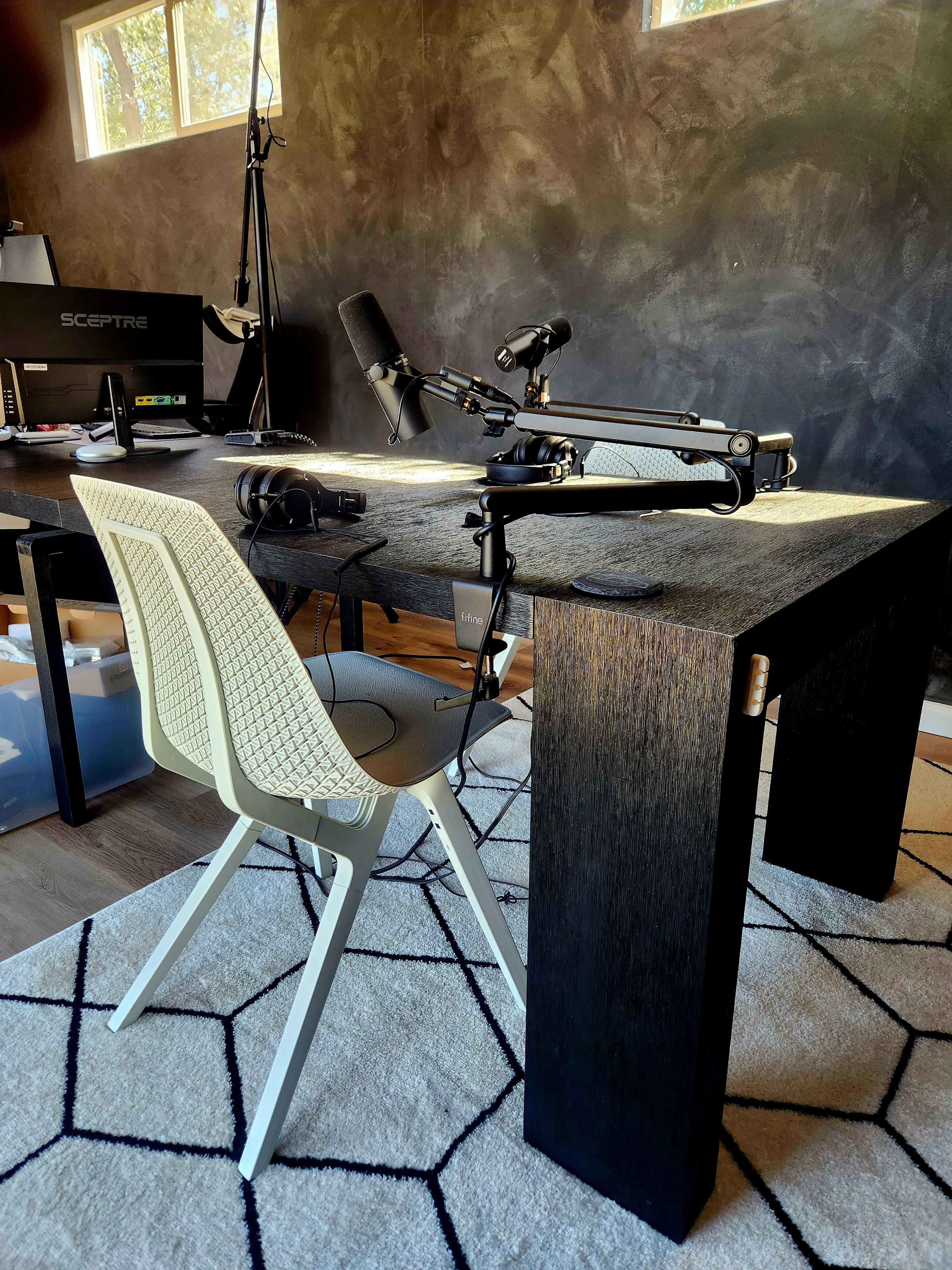 The image shows a modern podcasting setup with a table, a microphone on an adjustable arm, a chair, and a computer monitor in an office space.