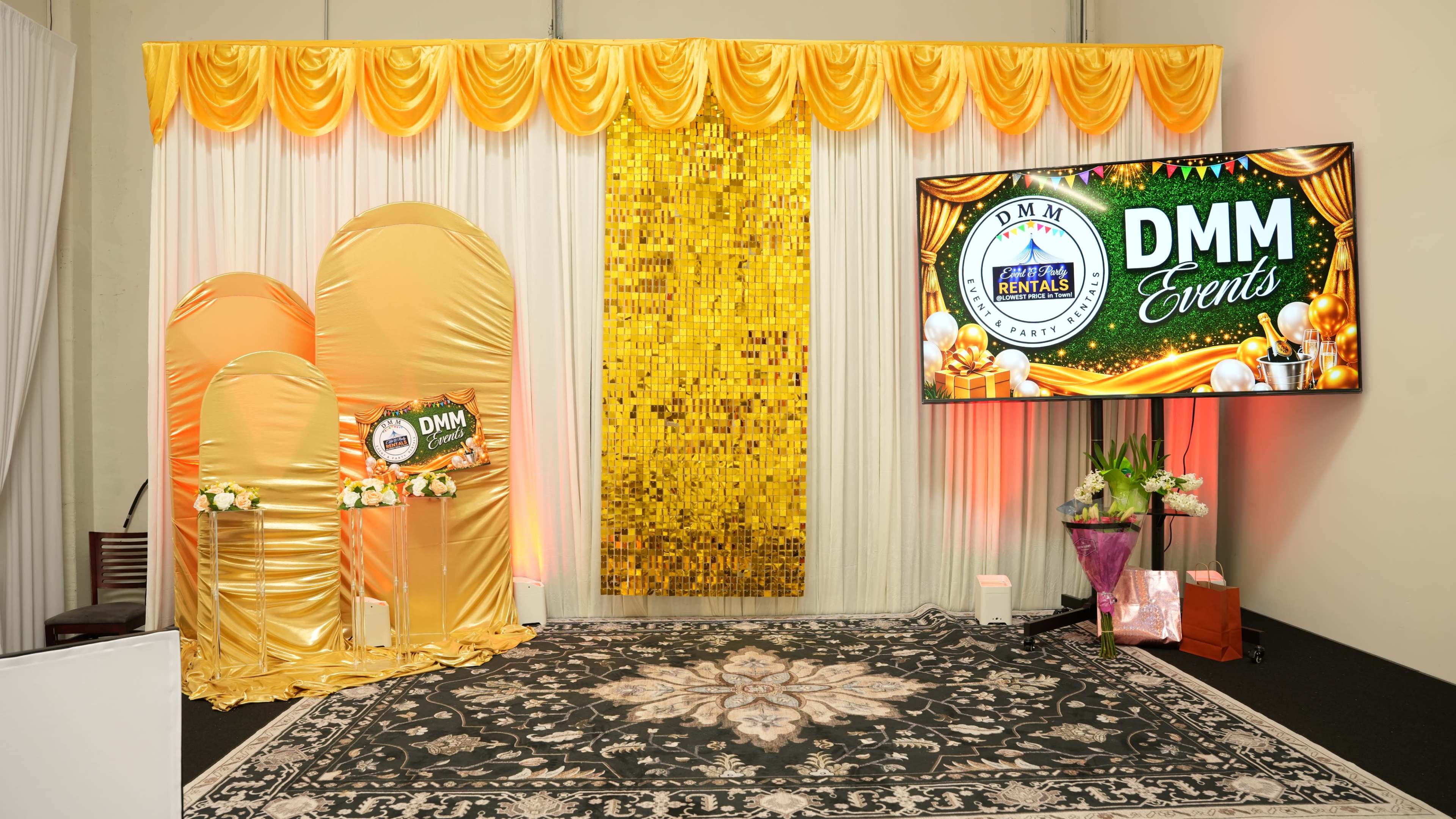 The image showcases a decorated event space featuring a golden backdrop, floral arrangements, and a large screen displaying "DMM Events."