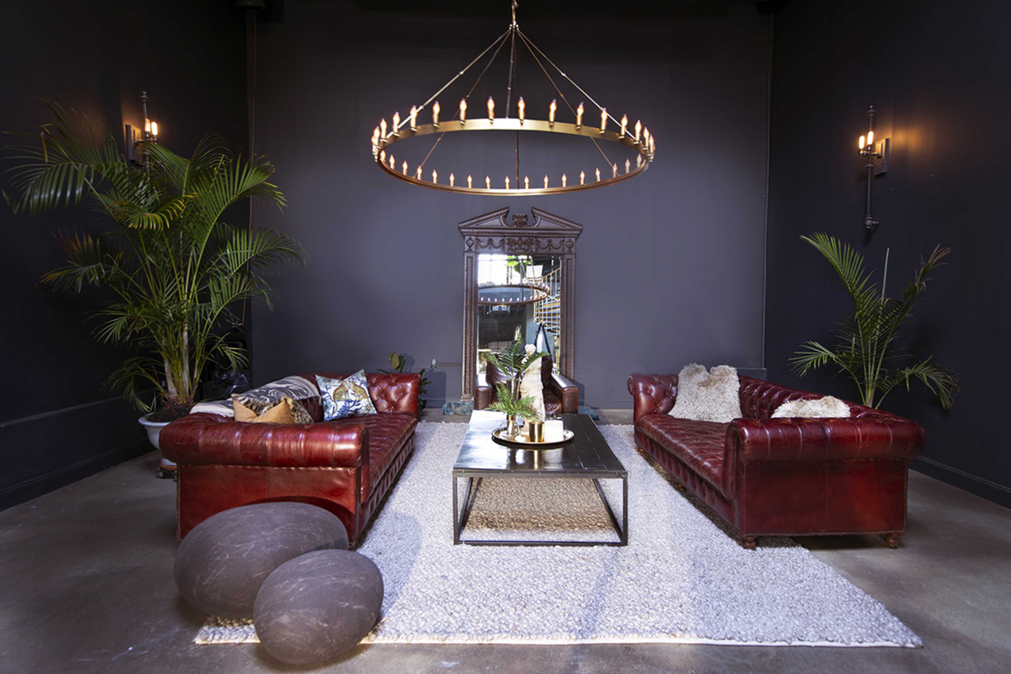 The image features a dark room with two plush red leather sofas facing each other, a large mirror, a circular chandelier, and potted plants.