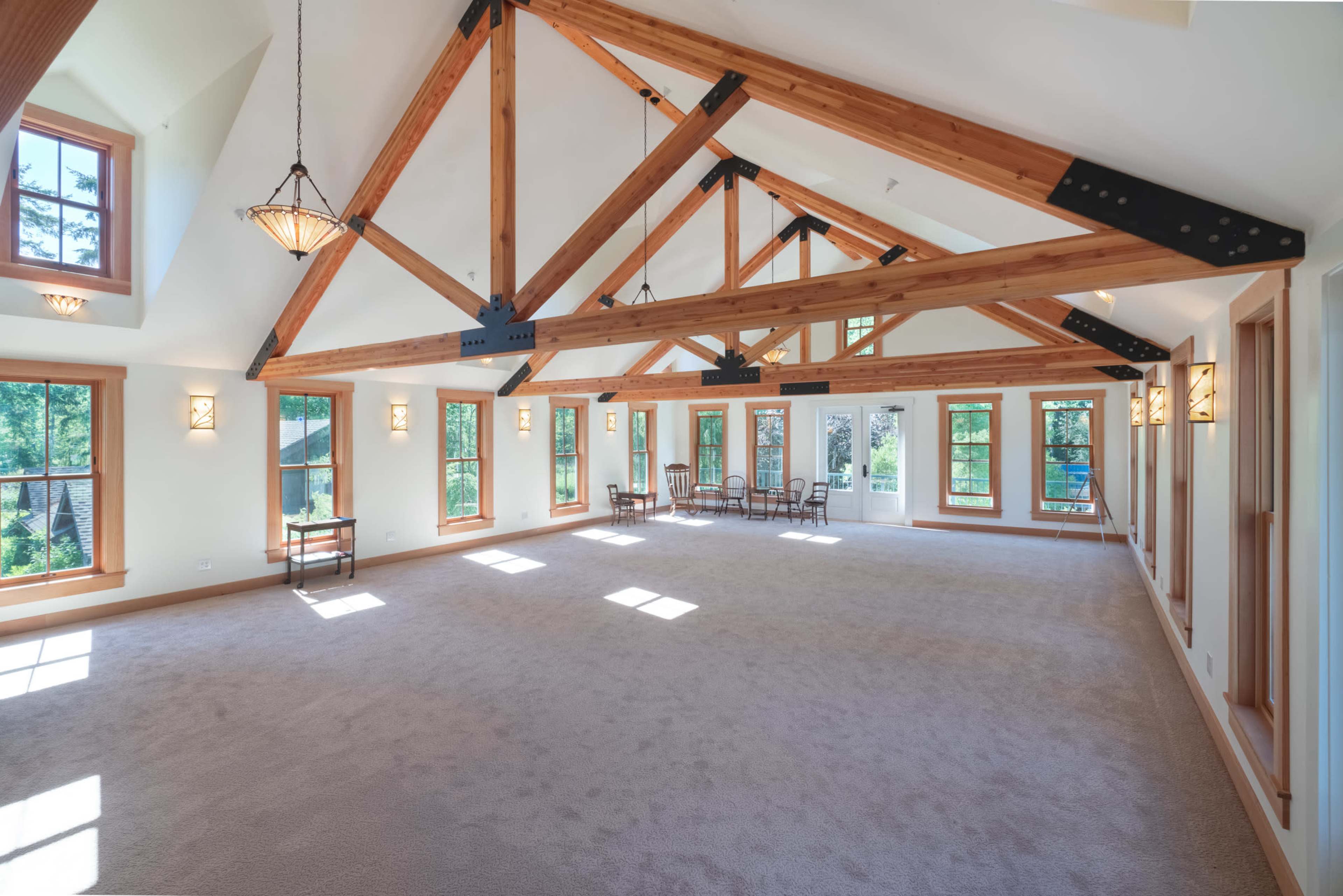 The spacious room features high, exposed wooden beams and multiple windows allowing natural light to fill the carpeted space.