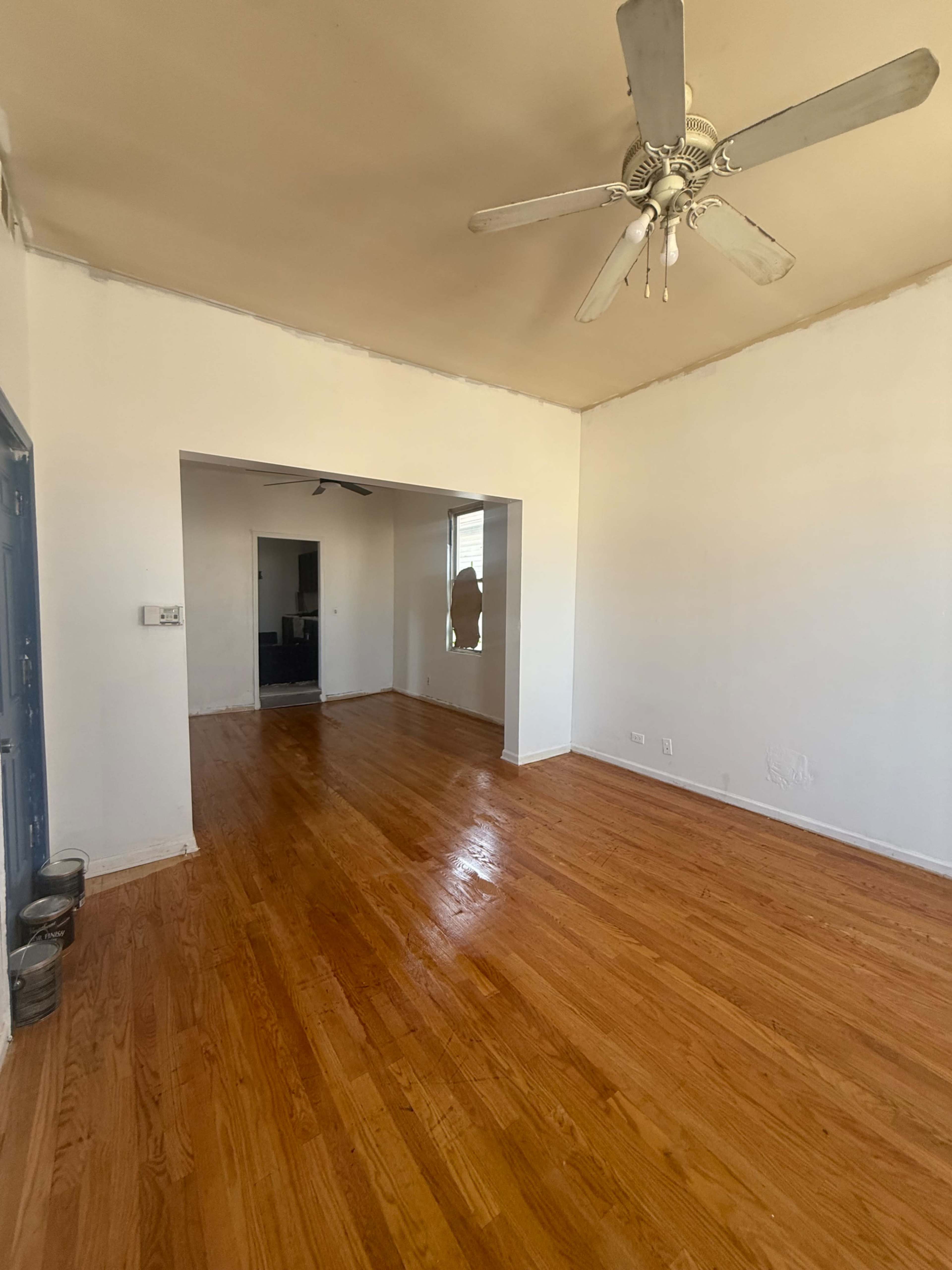 The image shows a spacious room with hardwood floors, a ceiling fan, and an opening leading to another space.