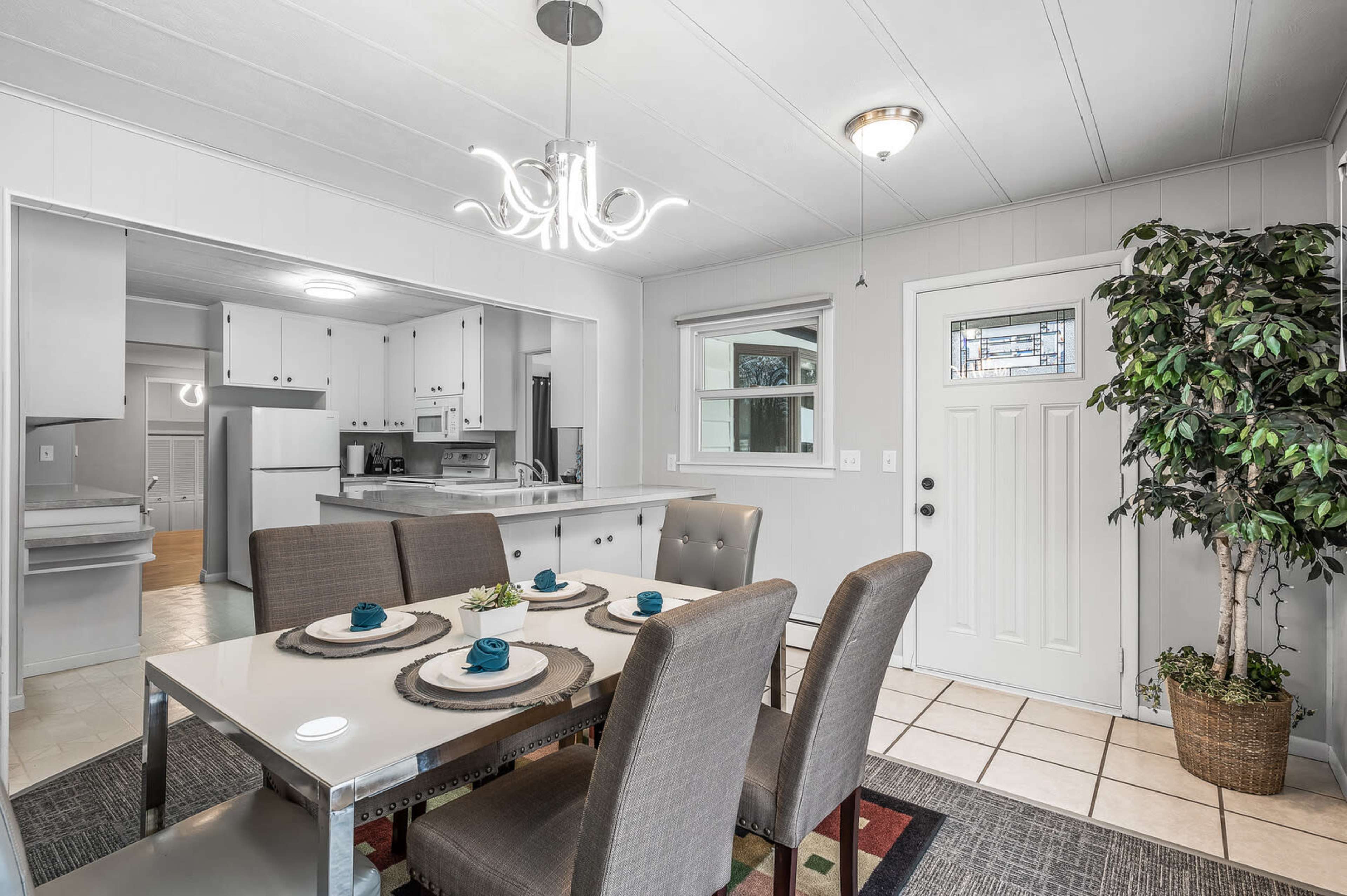 A modern dining area featuring a table with gray upholstered chairs, a chandelier overhead, and a view into an adjacent kitchen.
