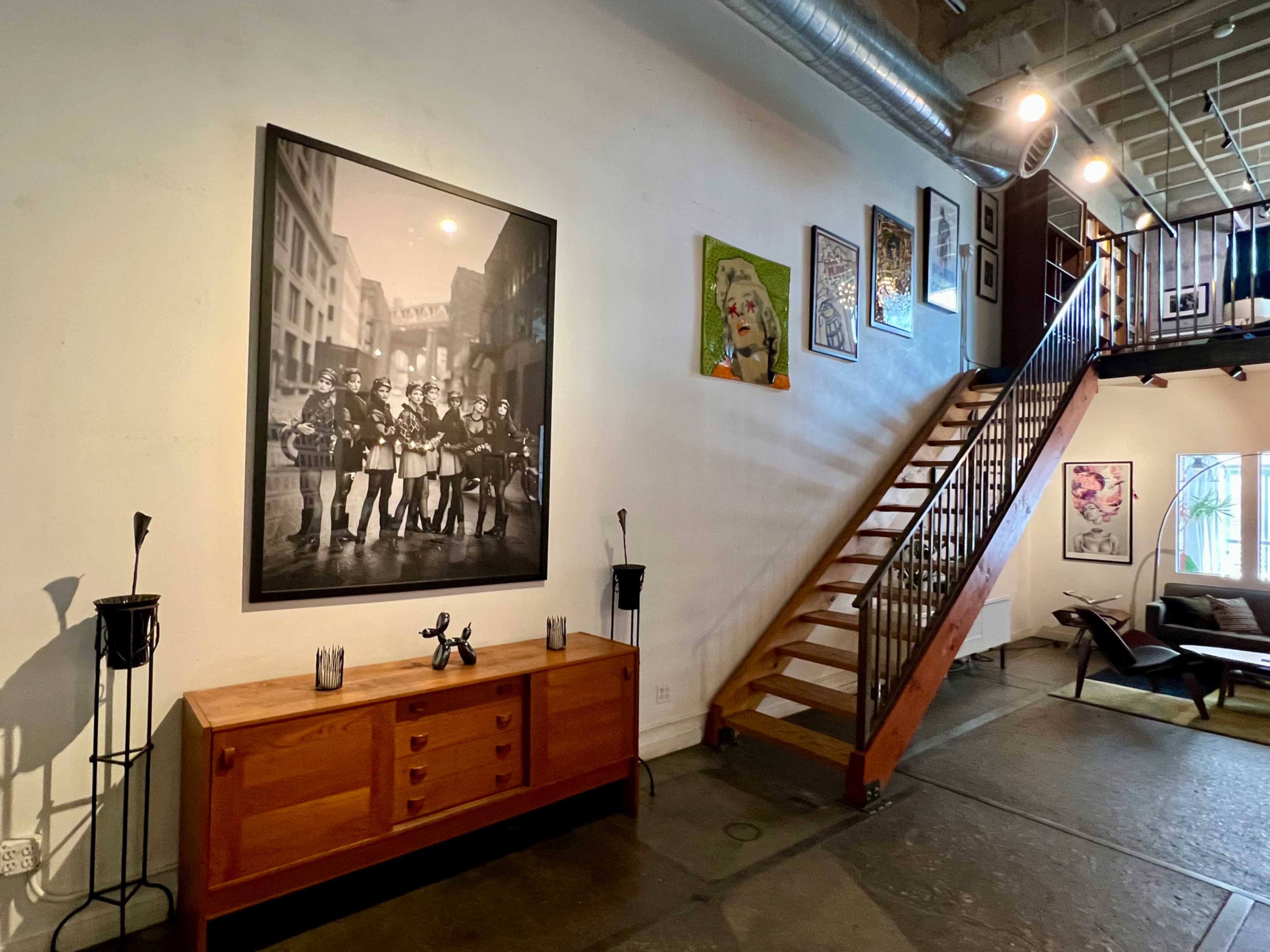 The image features a modern interior with a wooden staircase, a large black-and-white photograph hanging on the wall, and various artworks displayed alongside a wooden sideboard.
