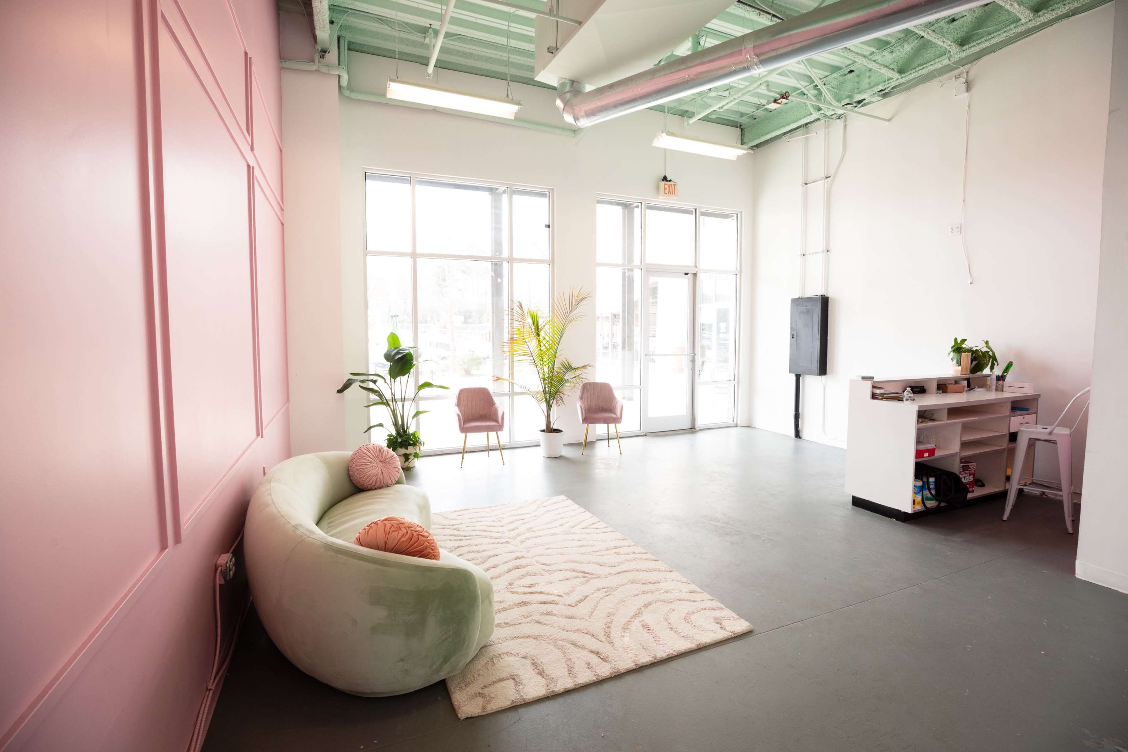 A bright, minimalist interior space featuring a curved green sofa, a light rug, two pink chairs, and several plants by a large window.