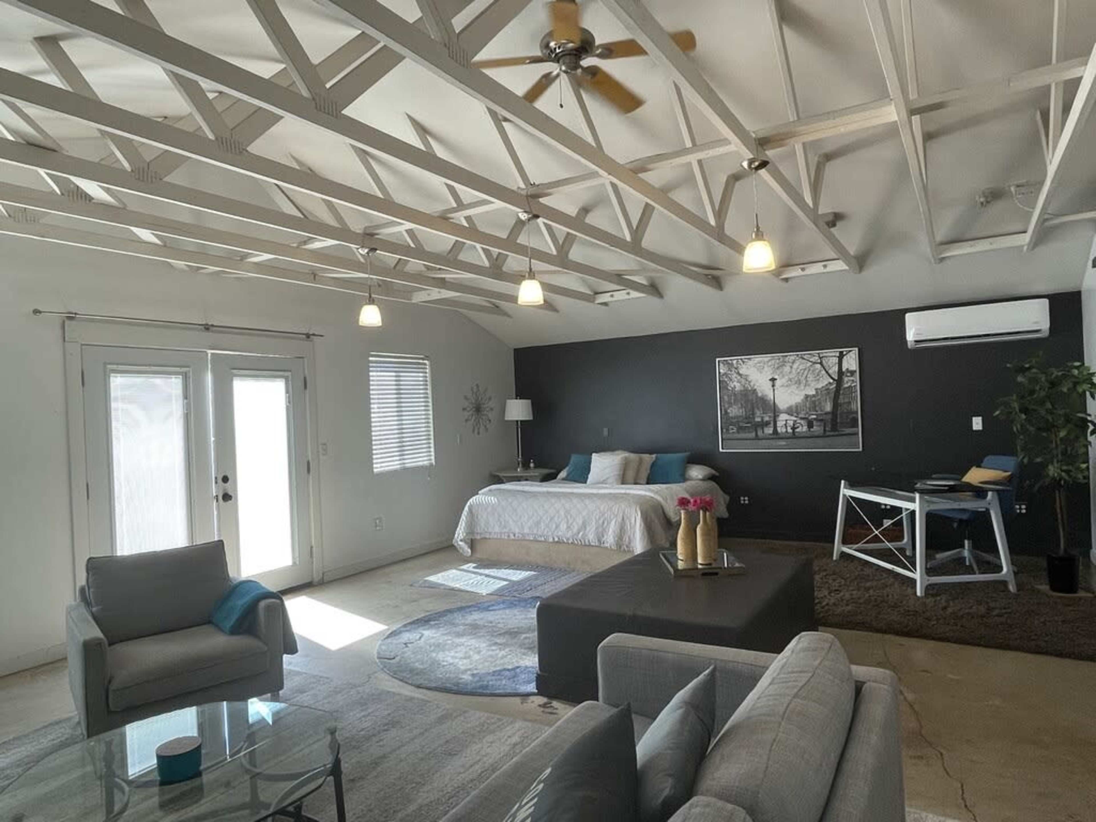 A bright, open living space featuring a bed, a desk, and a seating area beneath a vaulted ceiling with exposed beams.
