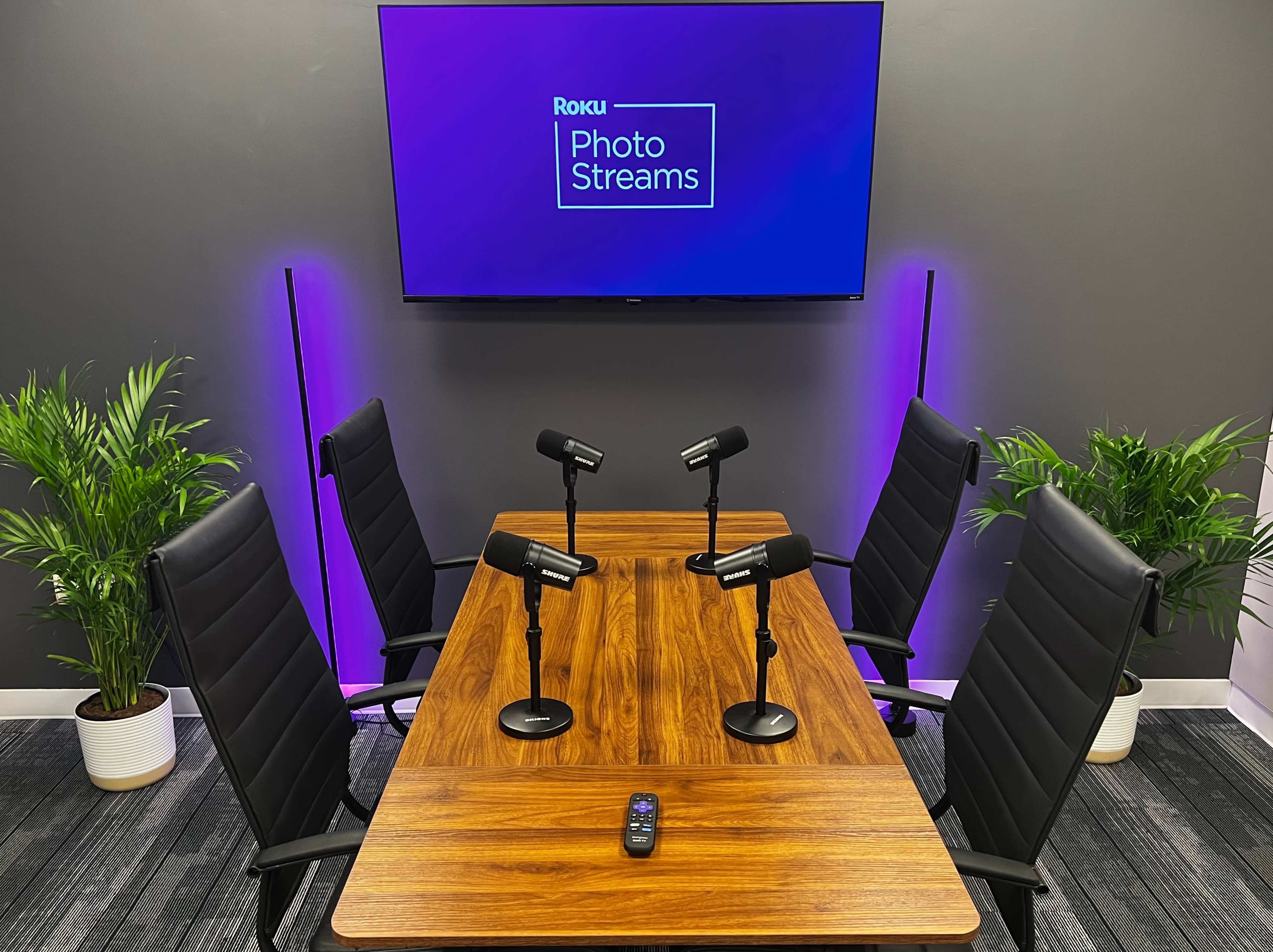 A meeting room is set up with a wooden table, four black chairs, and four microphones, featuring a Roku screen that says "Photo Streams" in the background.