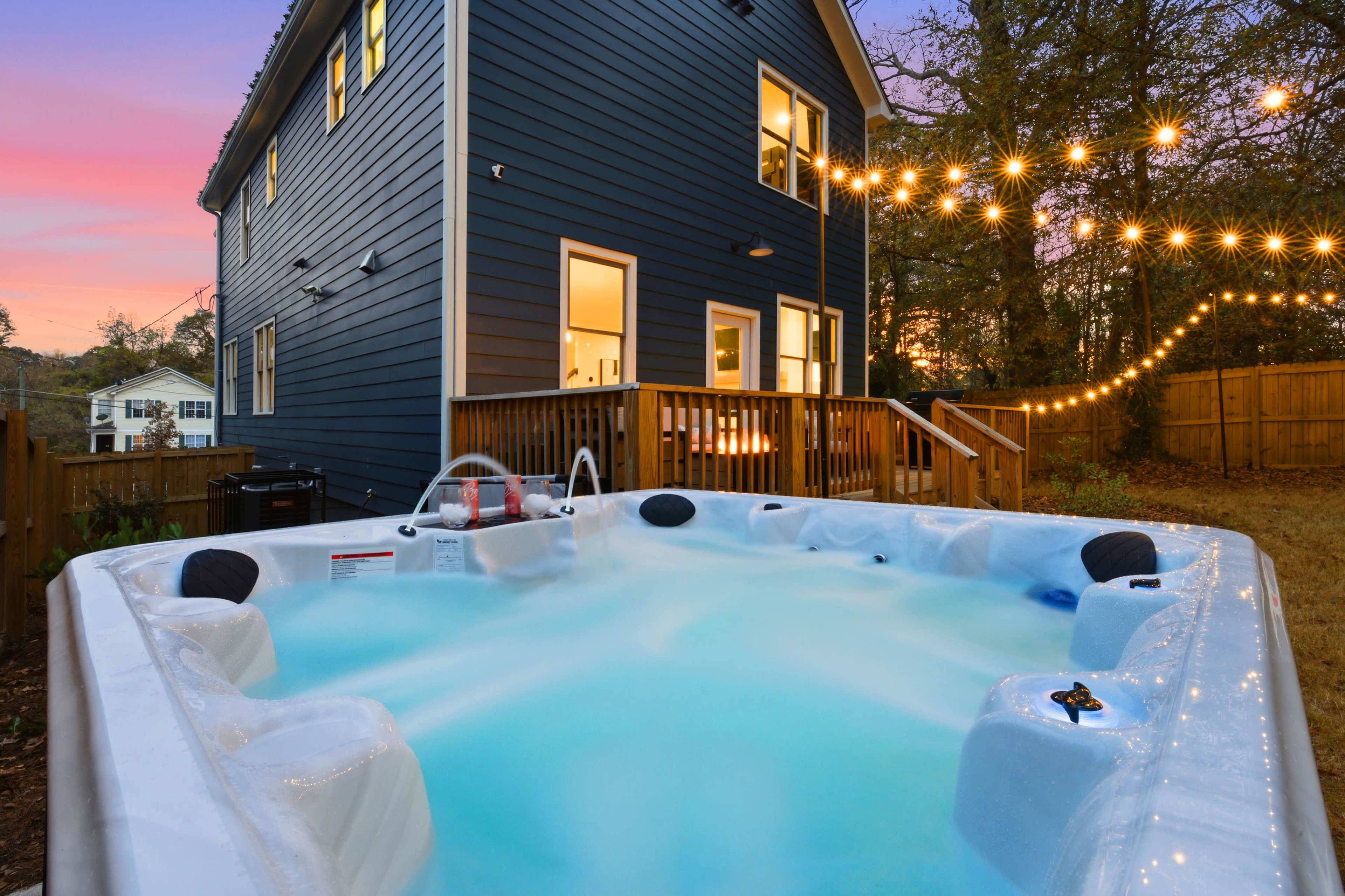 A hot tub sits in a backyard next to a two-story blue house, illuminated by string lights as dusk settles in.