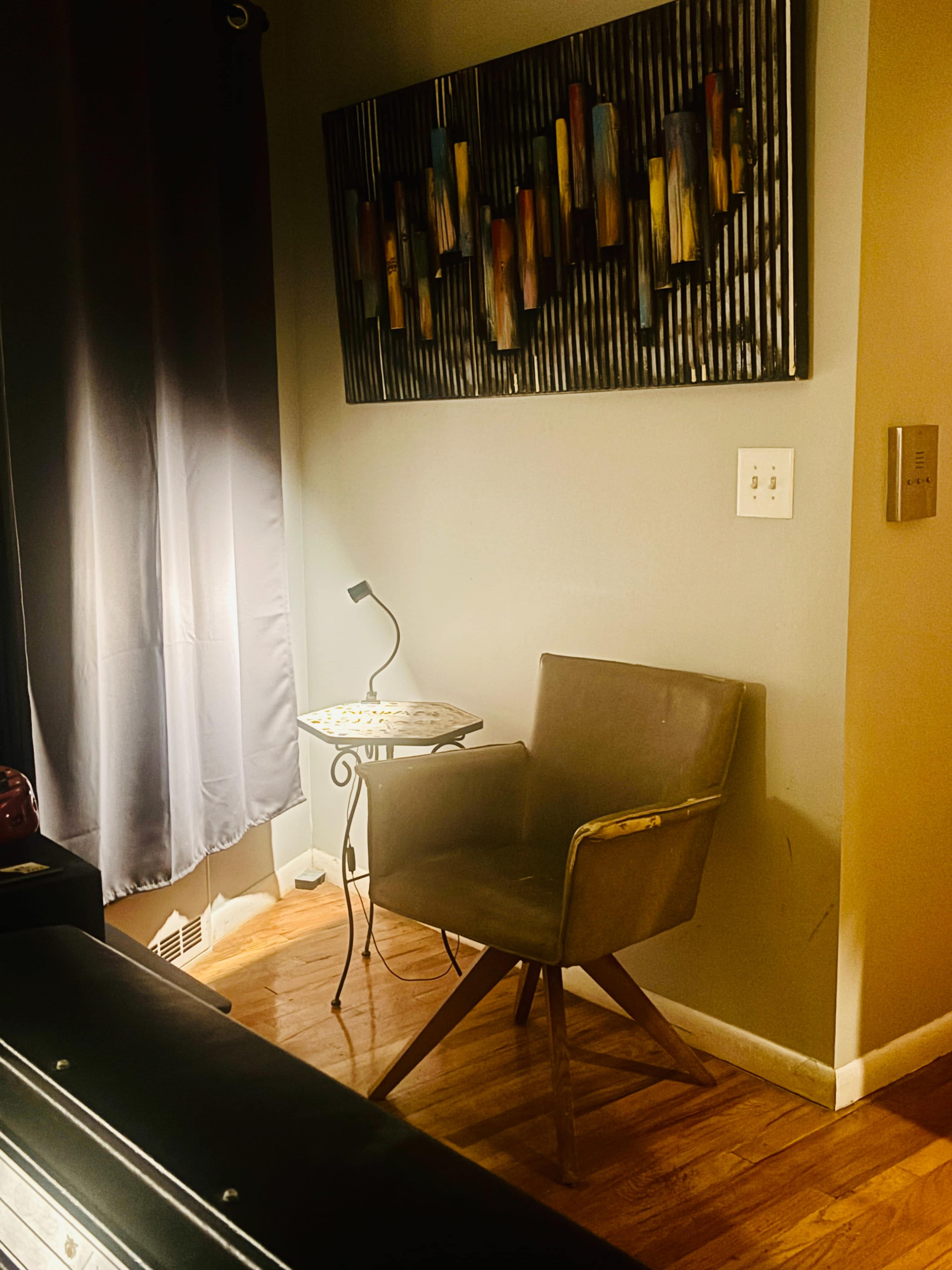 A brown armchair is positioned beside a small table under a wall art piece, with a lamp illuminating the corner of a room.