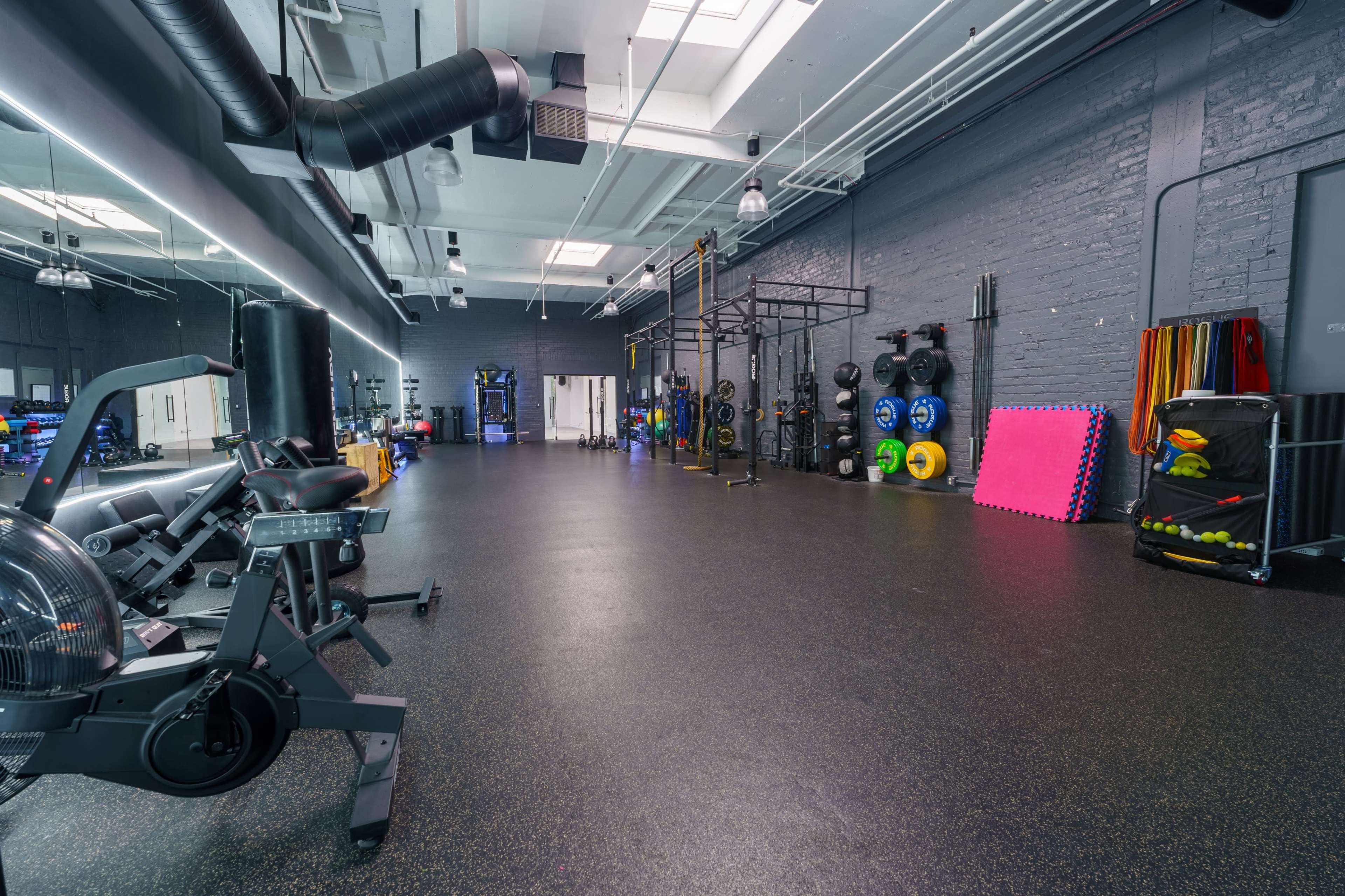 The image shows a spacious gym with various exercise equipment, including stationary bikes, weightlifting stations, and colorful weights along the walls.
