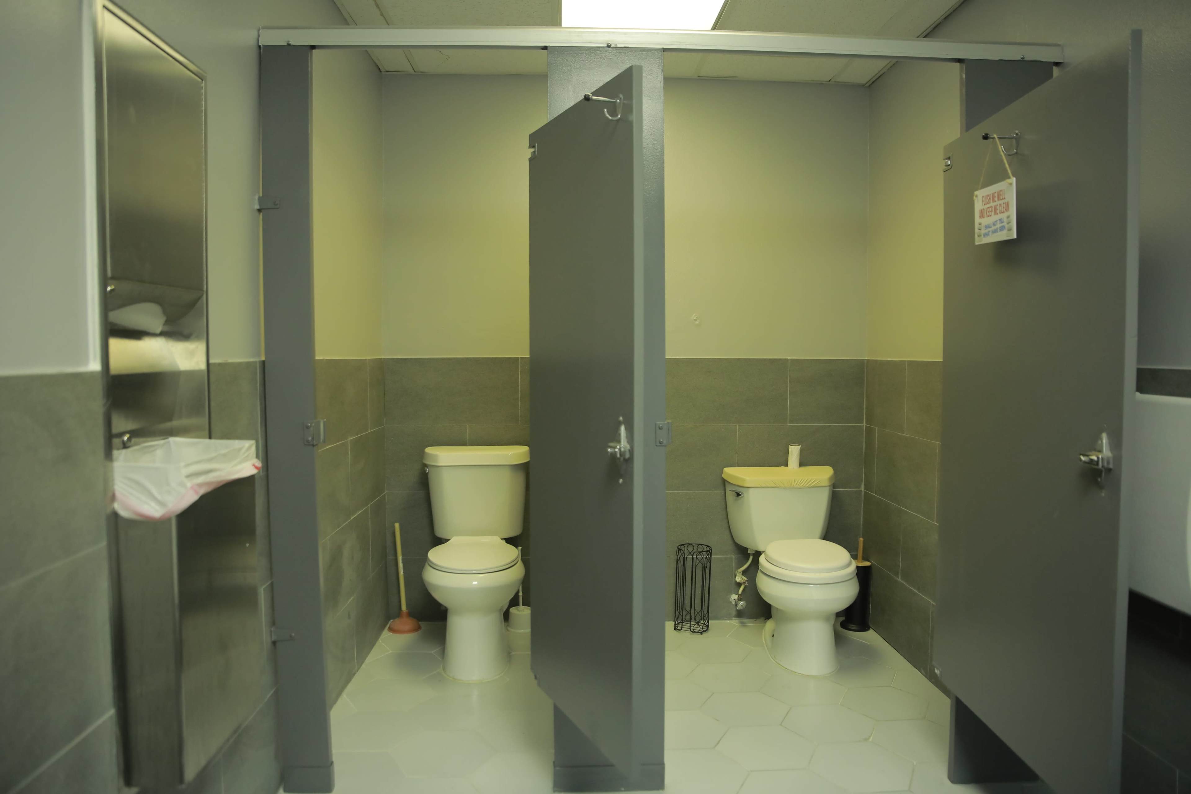 A public restroom with three toilet stalls, two of which are occupied, featuring gray partitions and tiled floors.