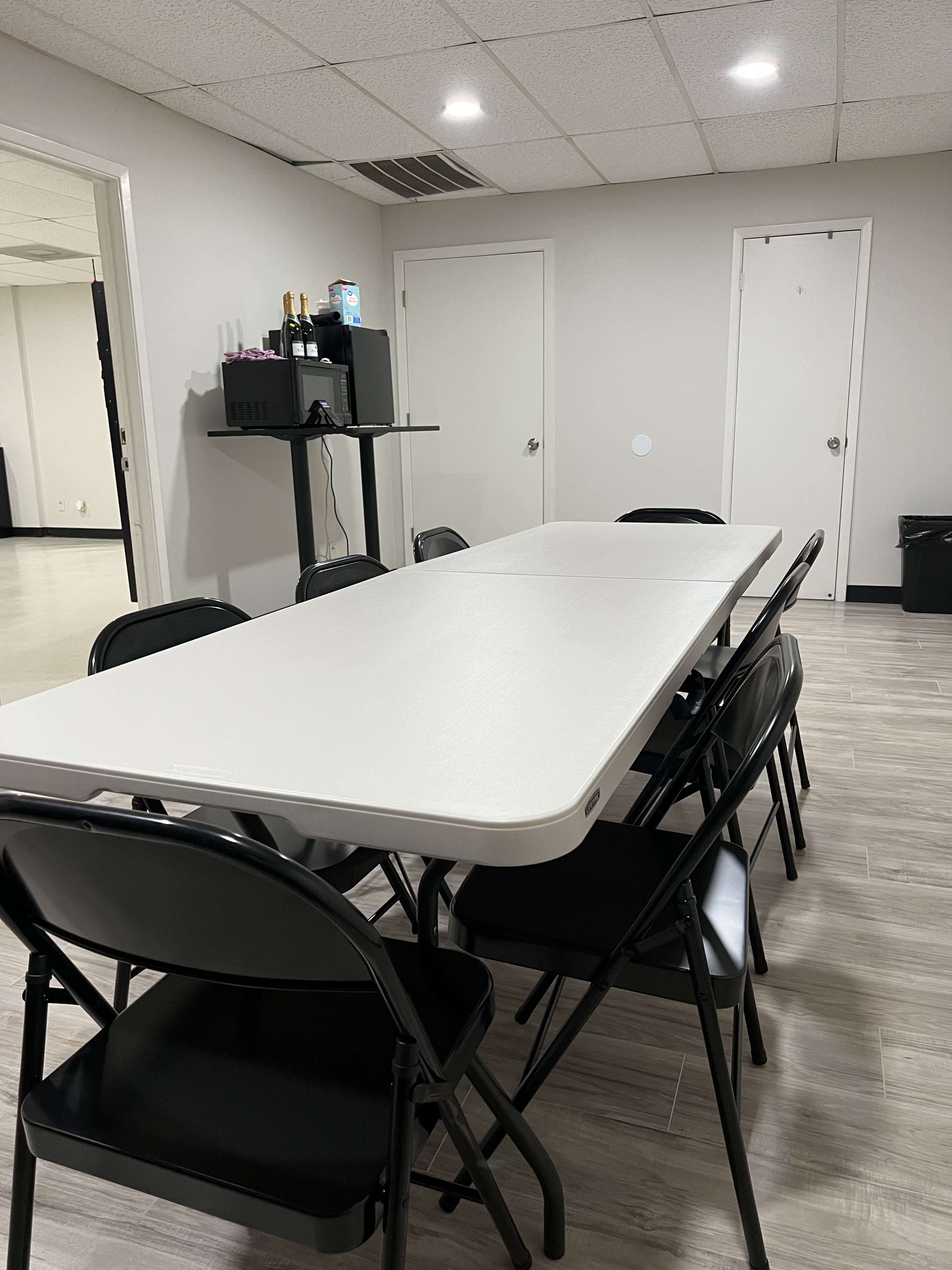 A rectangular table surrounded by black folding chairs is set up in a well-lit room with a door and a small shelf against the wall.