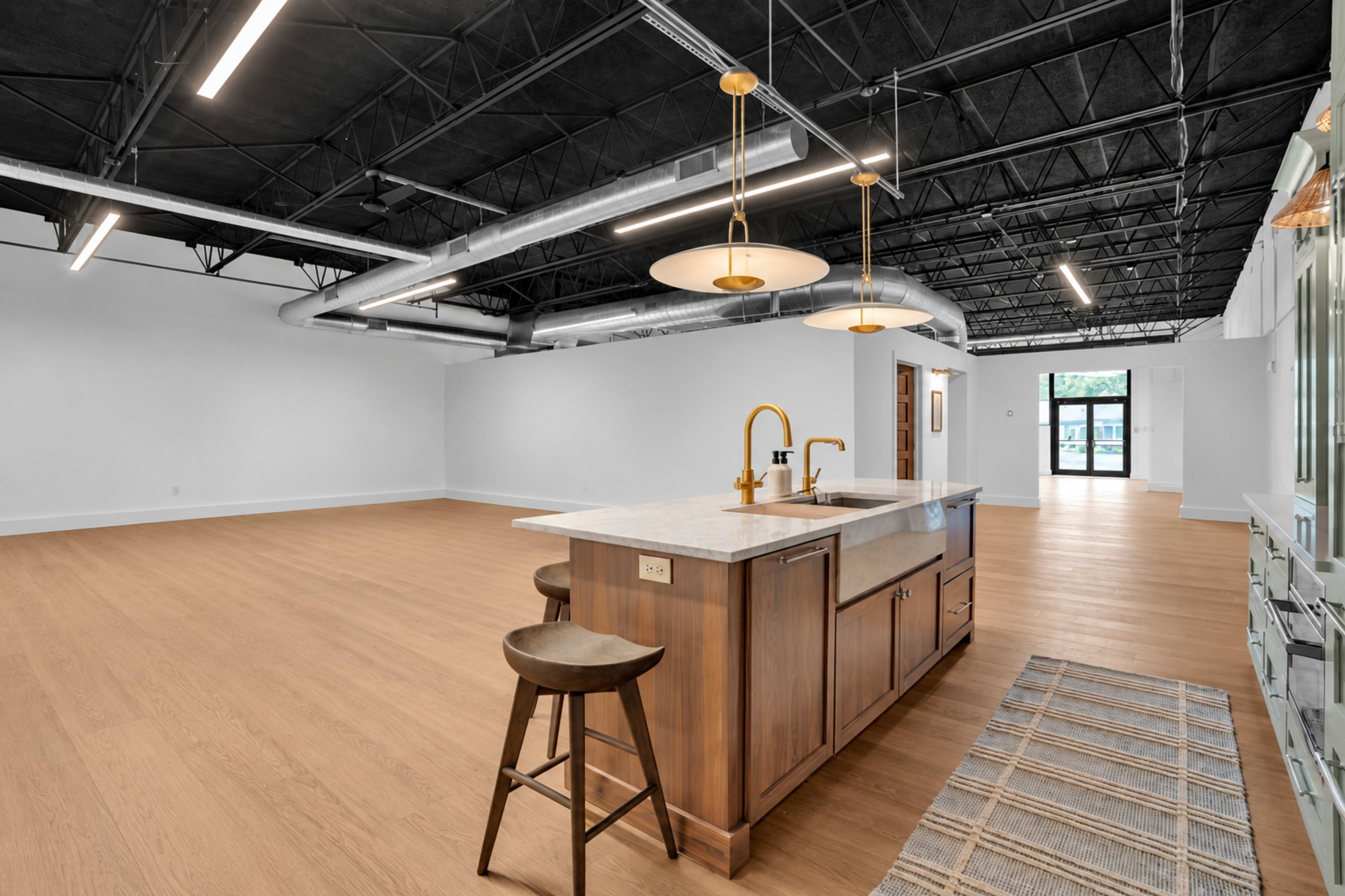 The image shows a spacious, open-concept room featuring a kitchen island with seating, beige wooden flooring, and large windows at one end.