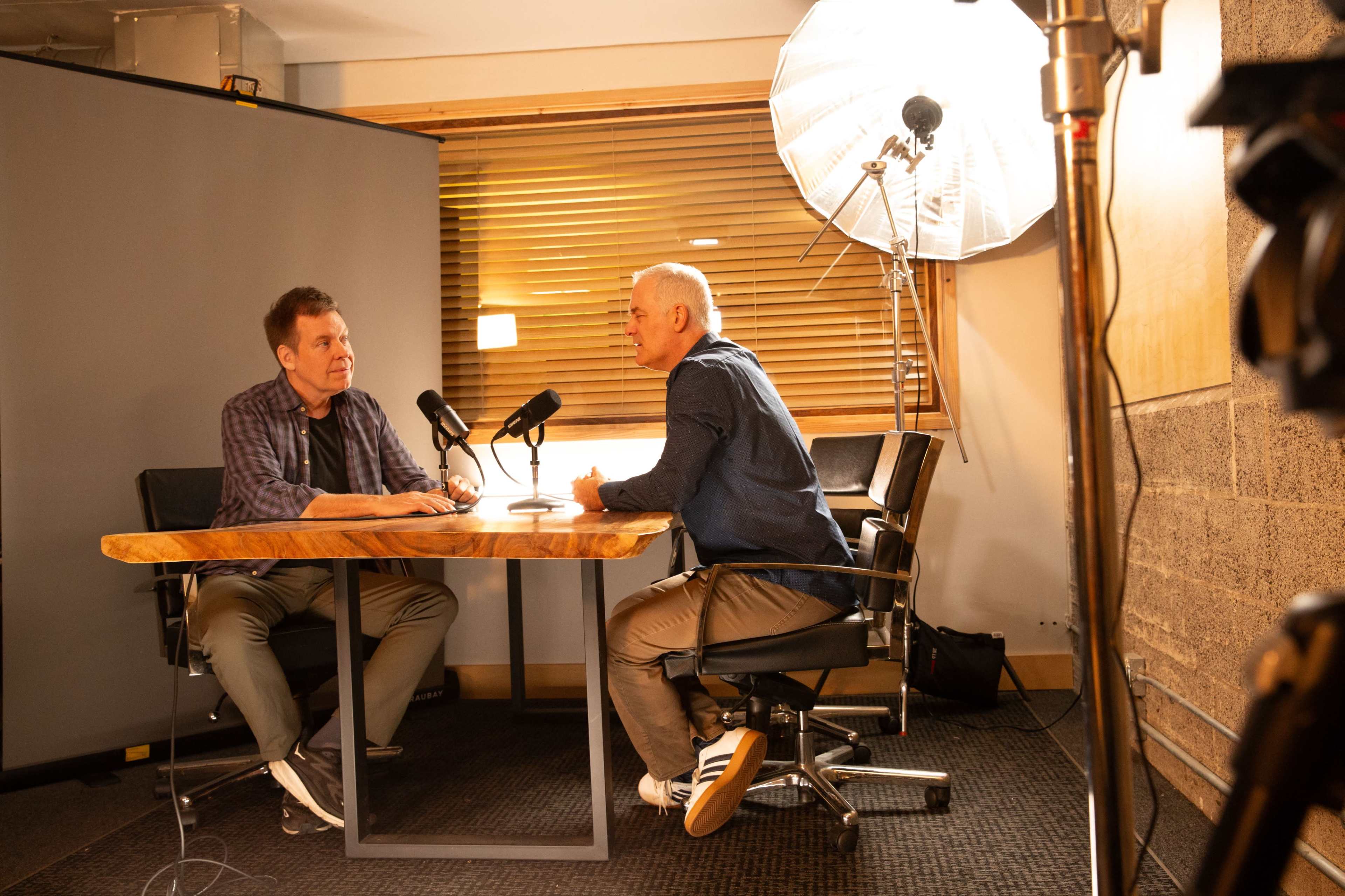 Two men are seated at a wooden table with microphones in front of them, engaged in conversation in a well-lit room.