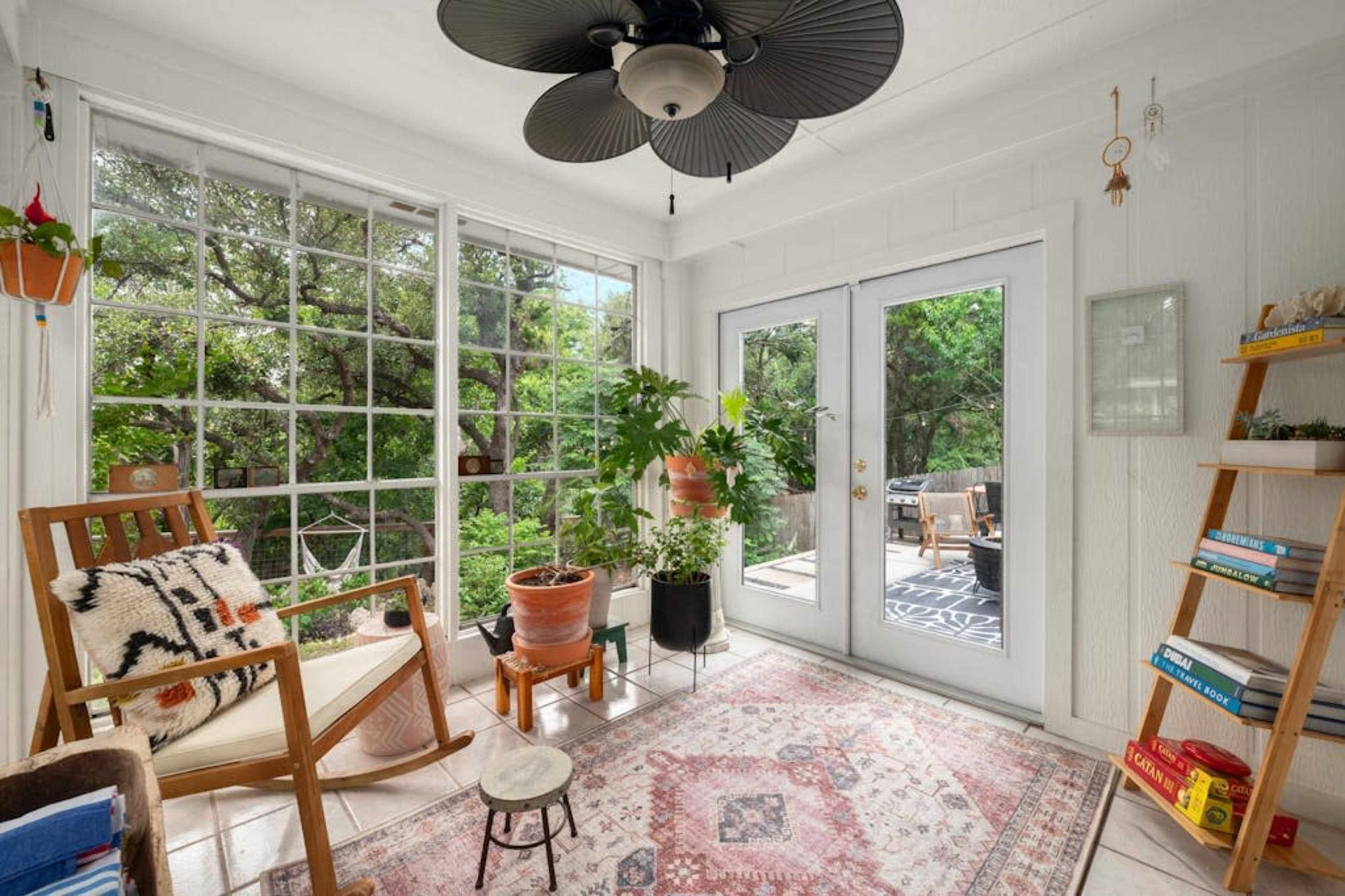 A bright sunroom features large windows, a wooden rocking chair, potted plants, and access to an outdoor seating area.